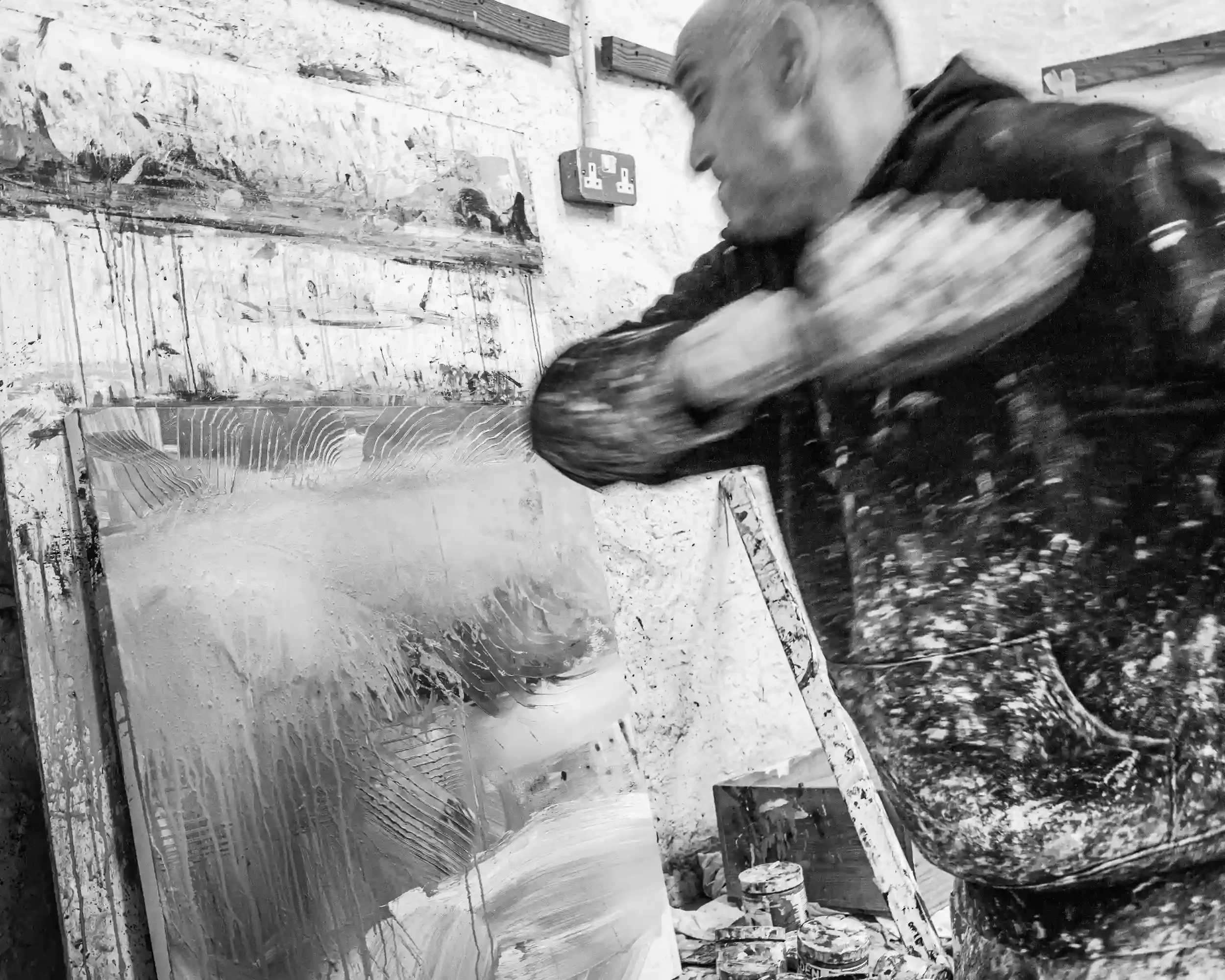 A dynamic black-and-white action shot of artist David Mankin in his Cornwall studio. His arm is a blur of motion as he sweeps a tool across a canvas, creating a series of textured, arched marks. He wears a paint-splattered hoodie, and the background 