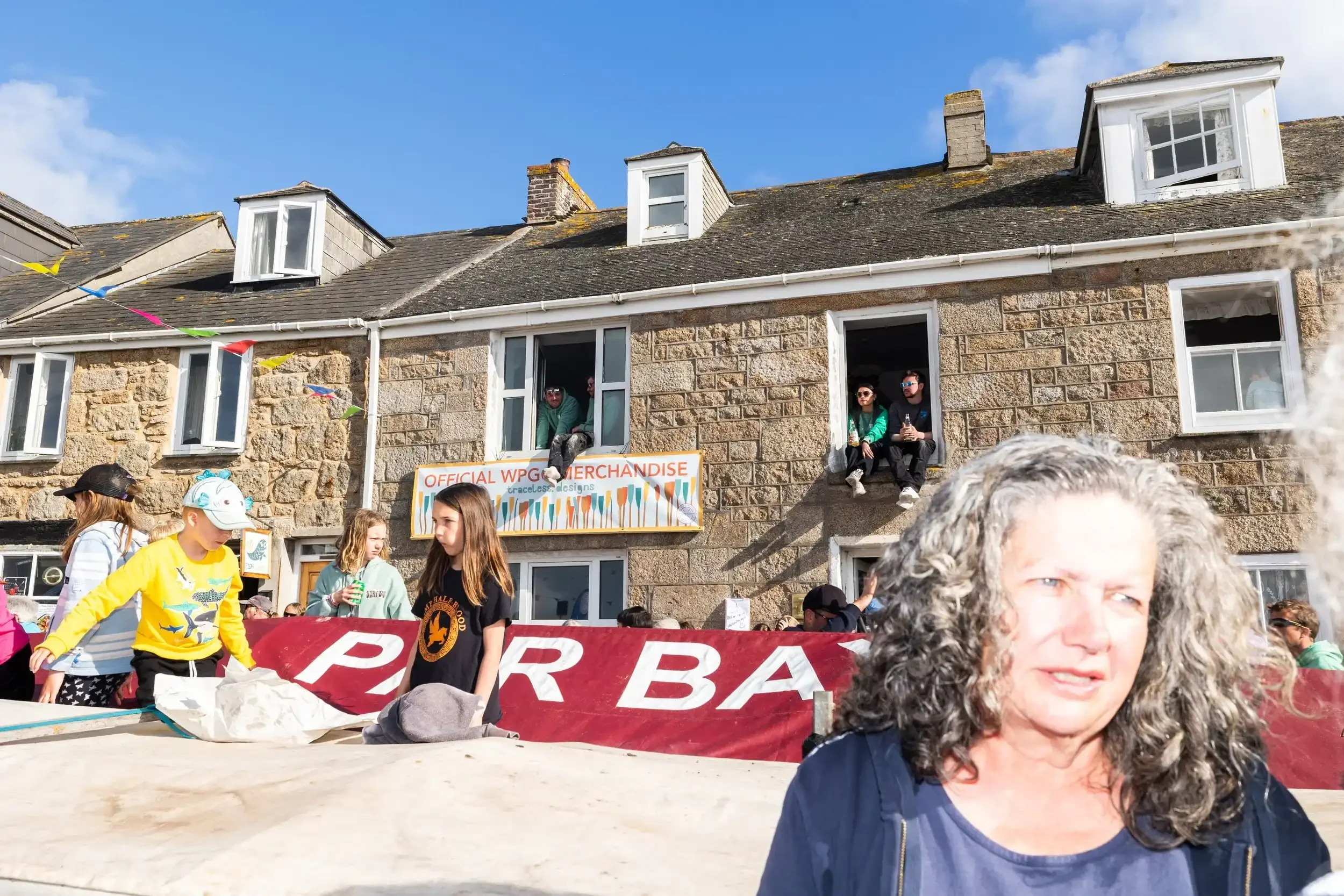 A wide-angle candid shot of a festive street celebration in the Isles of Scilly. In the foreground, a woman with long grey hair looks toward the side of the frame. Behind her, children play near a large boat covered with a maroon tarp. People are lea