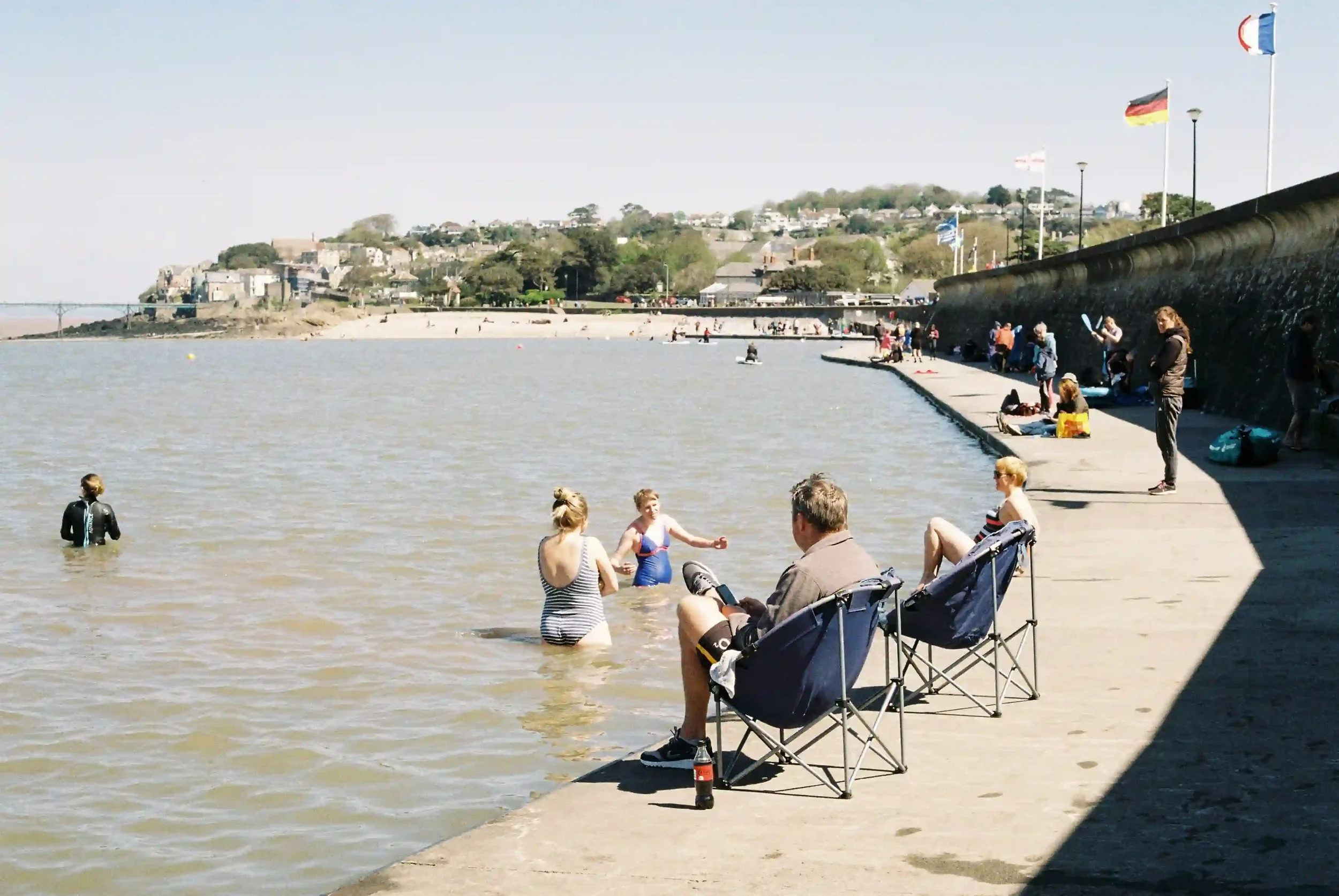 A wide shot of Clevedon Marine Lake on a clear day, with two women standing in the water, a man in a blue folding chair looking at his phone on the pier, and various flags flying along a high sea wall in the background, by Matthew Morgan.