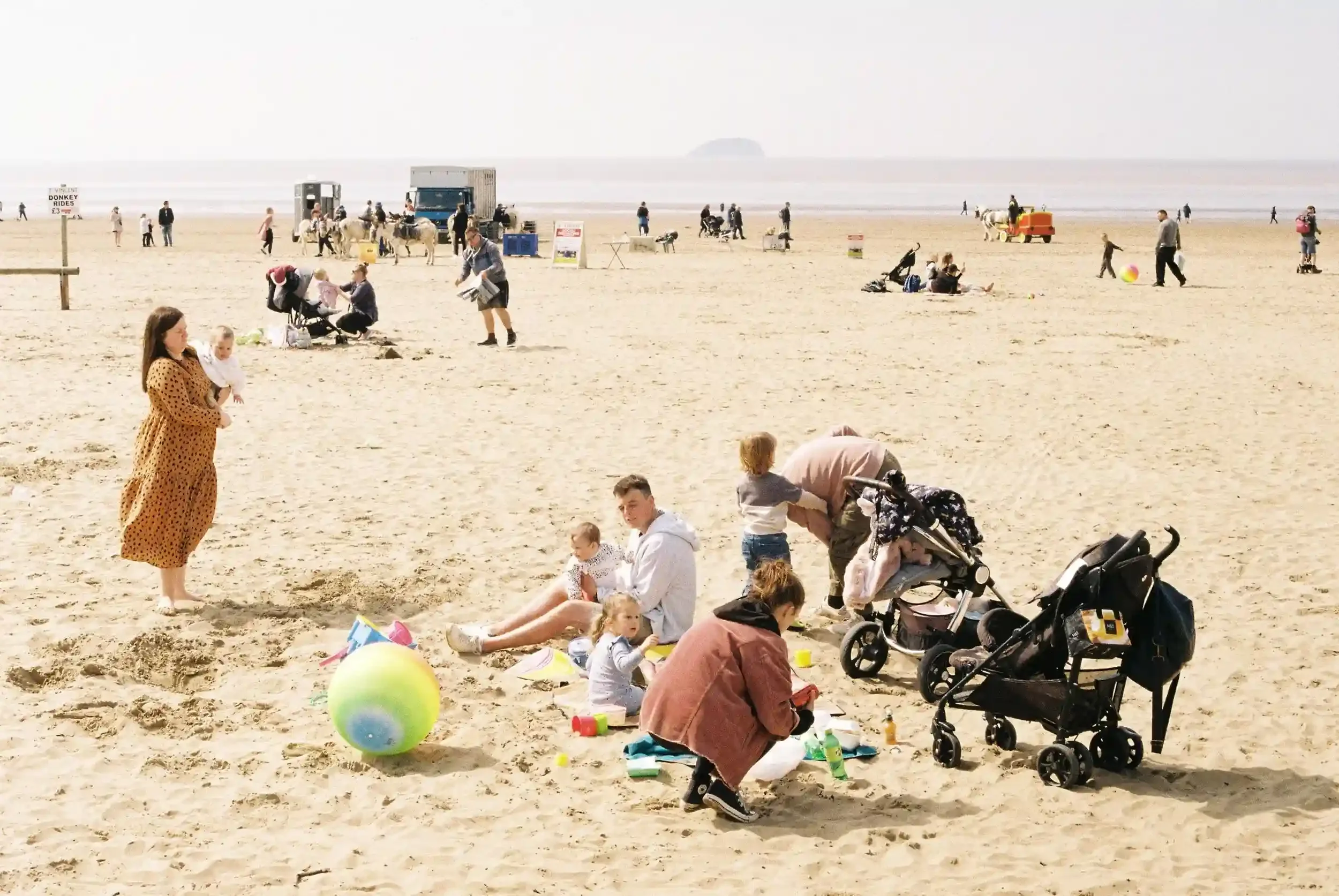 A busy, sun-bleached beach scene at Weston-super-Mare in May 2021, featuring a mother holding a baby and a family sitting on the sand with strollers and beach toys, by Matthew Morgan.