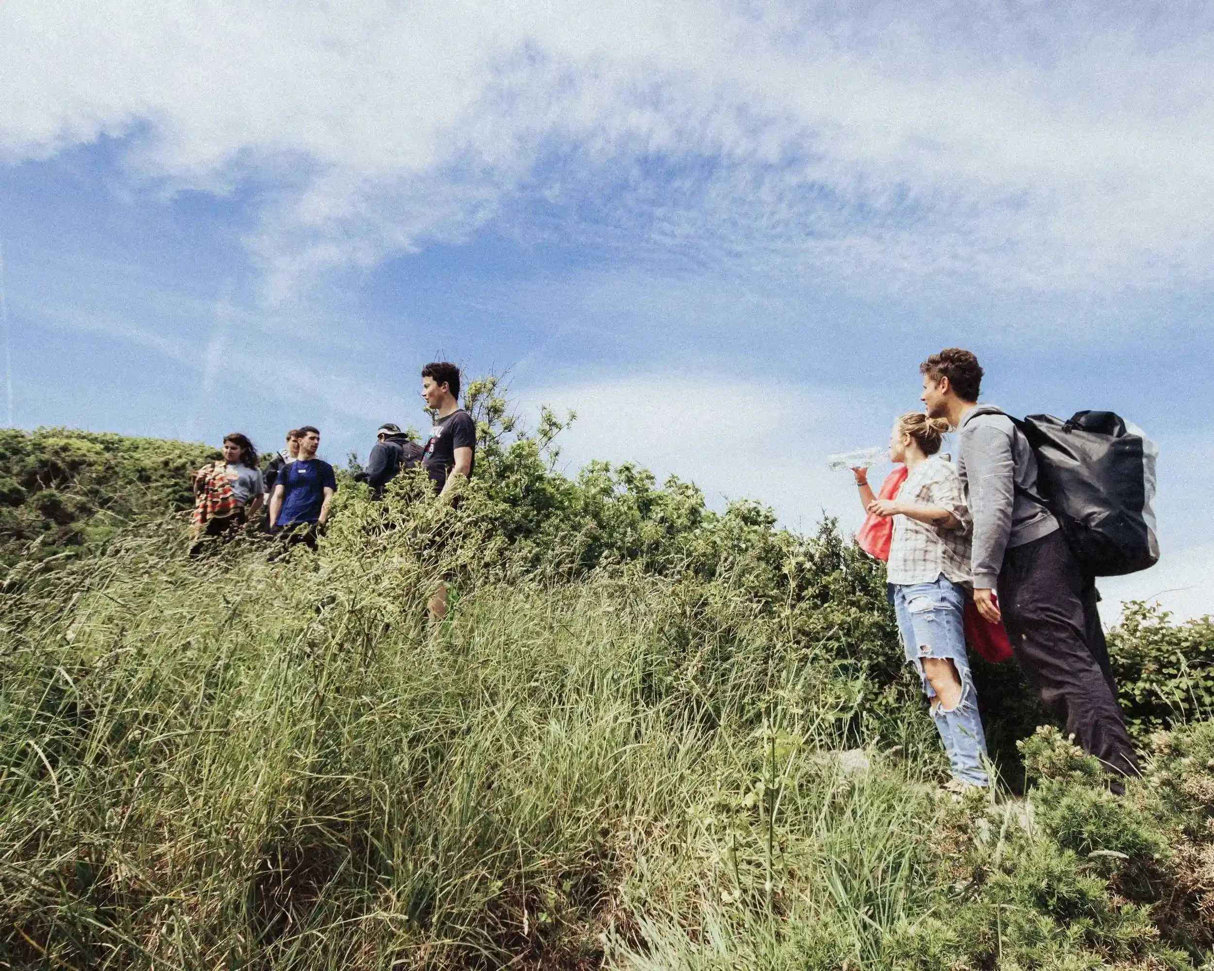 A wide-angle candid photograph of a group of people, including singer-songwriter Ned Holland, walking through tall grass on a coastal clifftop under a bright blue sky. Some crew members carry bags and water bottles, capturing a transition moment on t