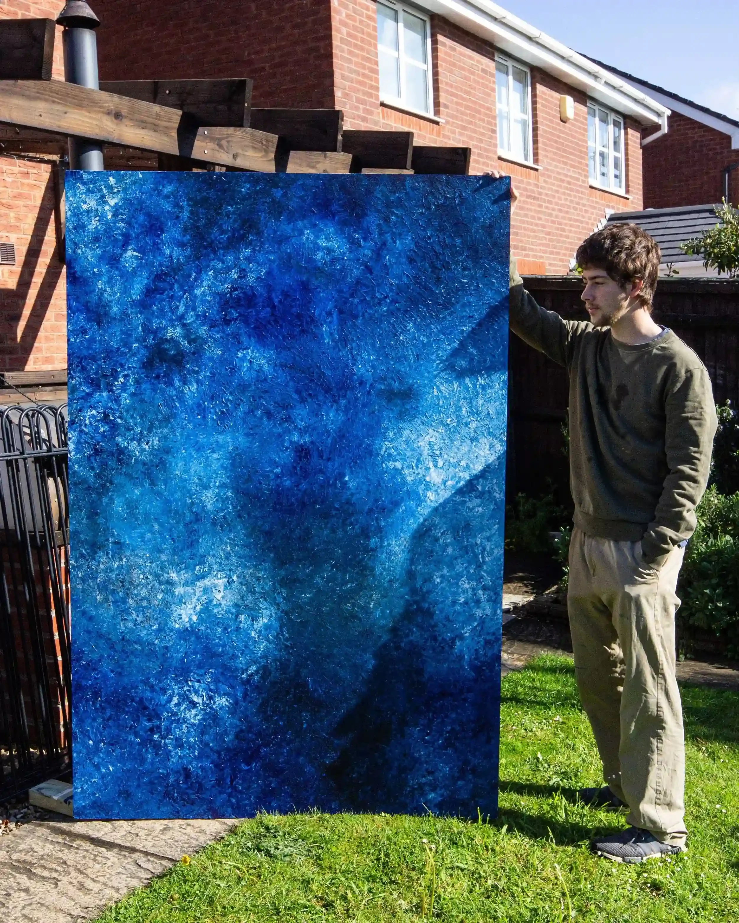 Artist Matthew Morgan standing in a grassy garden, holding a massive 6-foot by 4-foot vertical canvas covered in deep, textured blue and white abstract painting, with a brick house in the background.