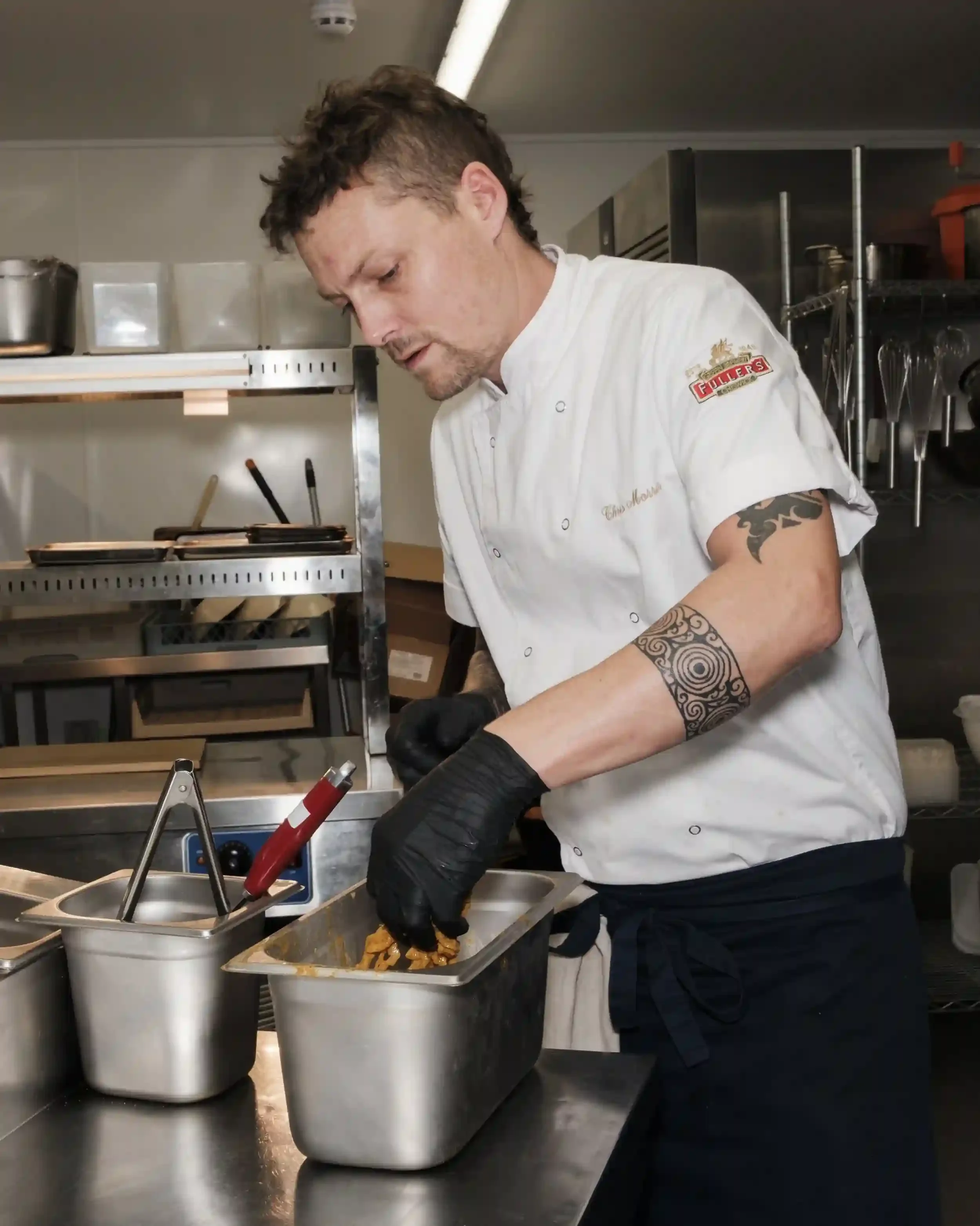 A candid photograph of the Chef working in the commercial kitchen of The Crown Inn, Lea. The chef, wearing a white short-sleeved jacket with a 'Fuller’s' logo and black apron, is focused on preparing food in a stainless steel container. His black-glo