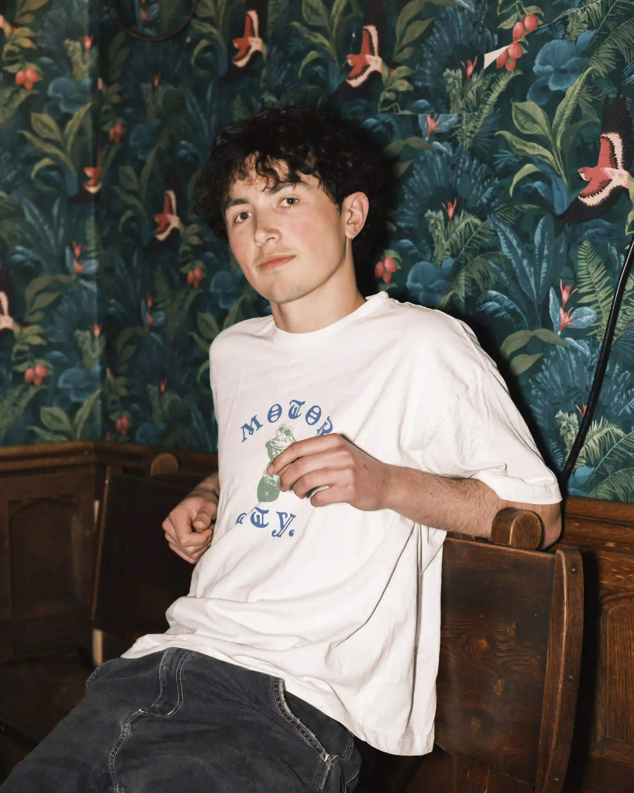 A clear, well-lit portrait of Ned Holland sitting on a wooden bench at The Troubadour, London. He is wearing a white 'Motor City' graphic t-shirt and dark jeans. The background features a distinctive teal wallpaper with large green leaves and orange 