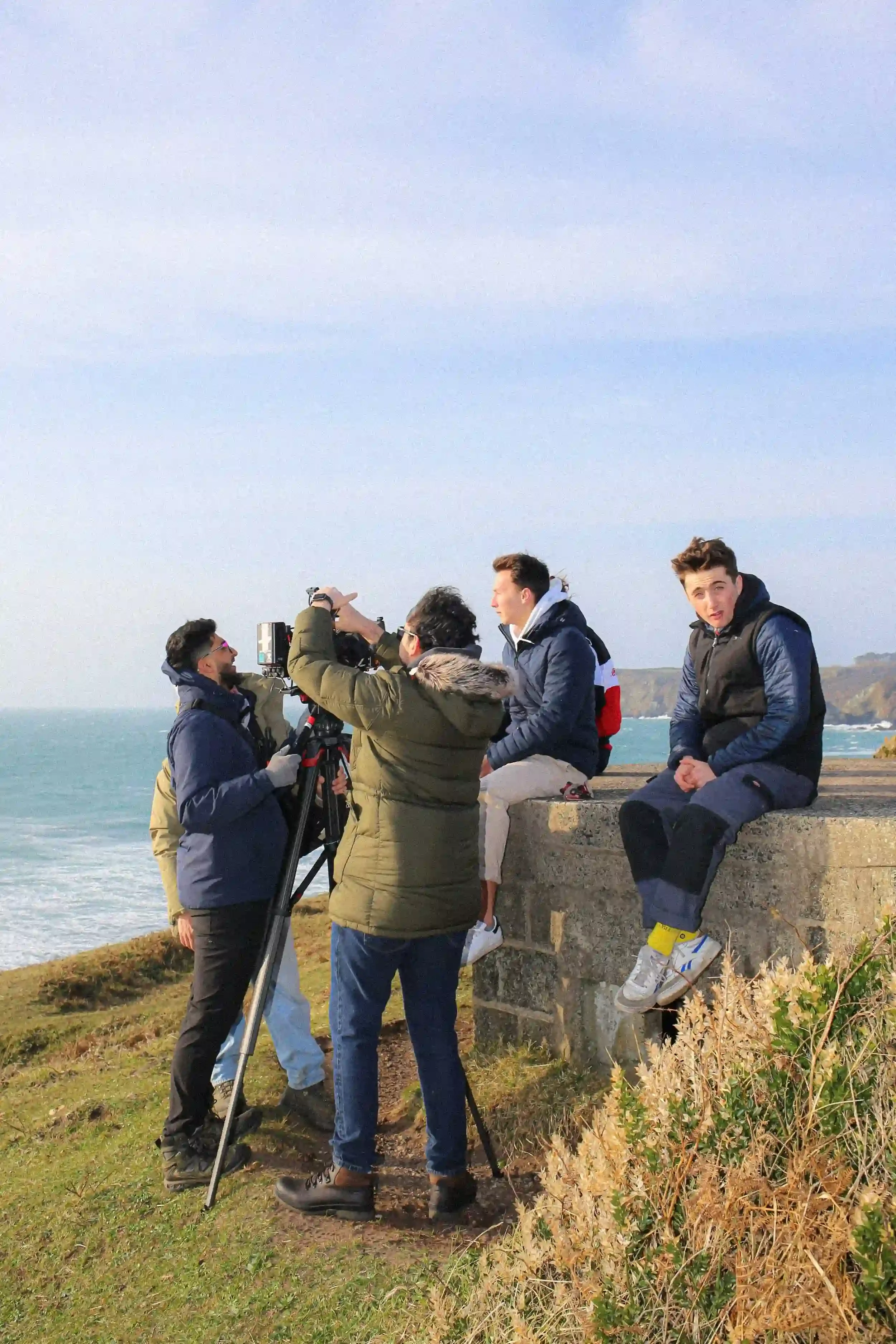 A behind-the-scenes colour photograph of a film crew working on a grassy cliffside overlooking a turquoise sea in Cornwall. Two crew members in warm jackets operate a professional cinema camera mounted on a large tripod, while two actors sit on a sto