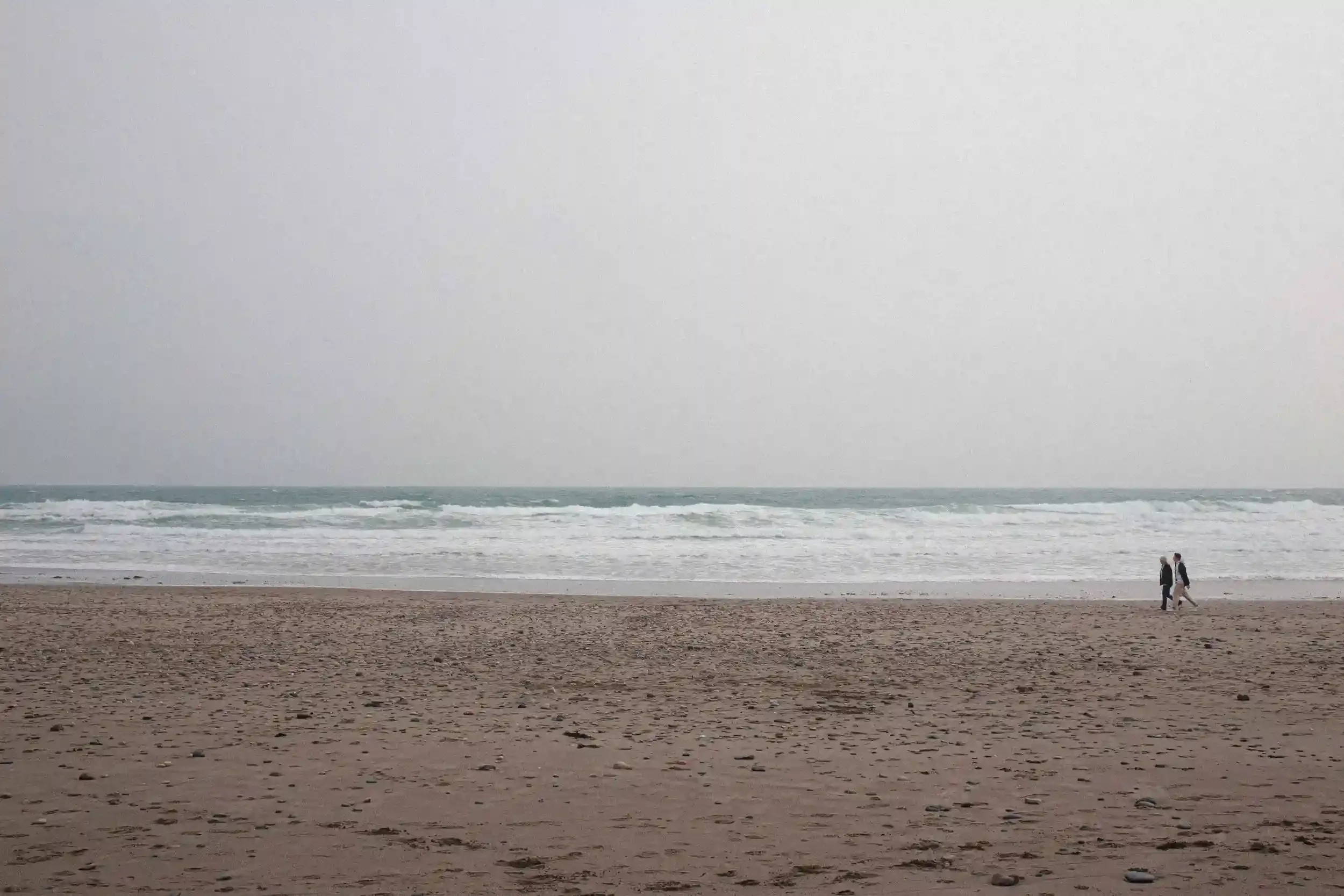 A wide-angle colour unit still from the film 'Lighter' showing an expansive sandy beach and a rough, white-capped sea under a heavy, overcast sky. On the far right of the frame, two small figures of a man and a woman walk along the wet sand. The mini