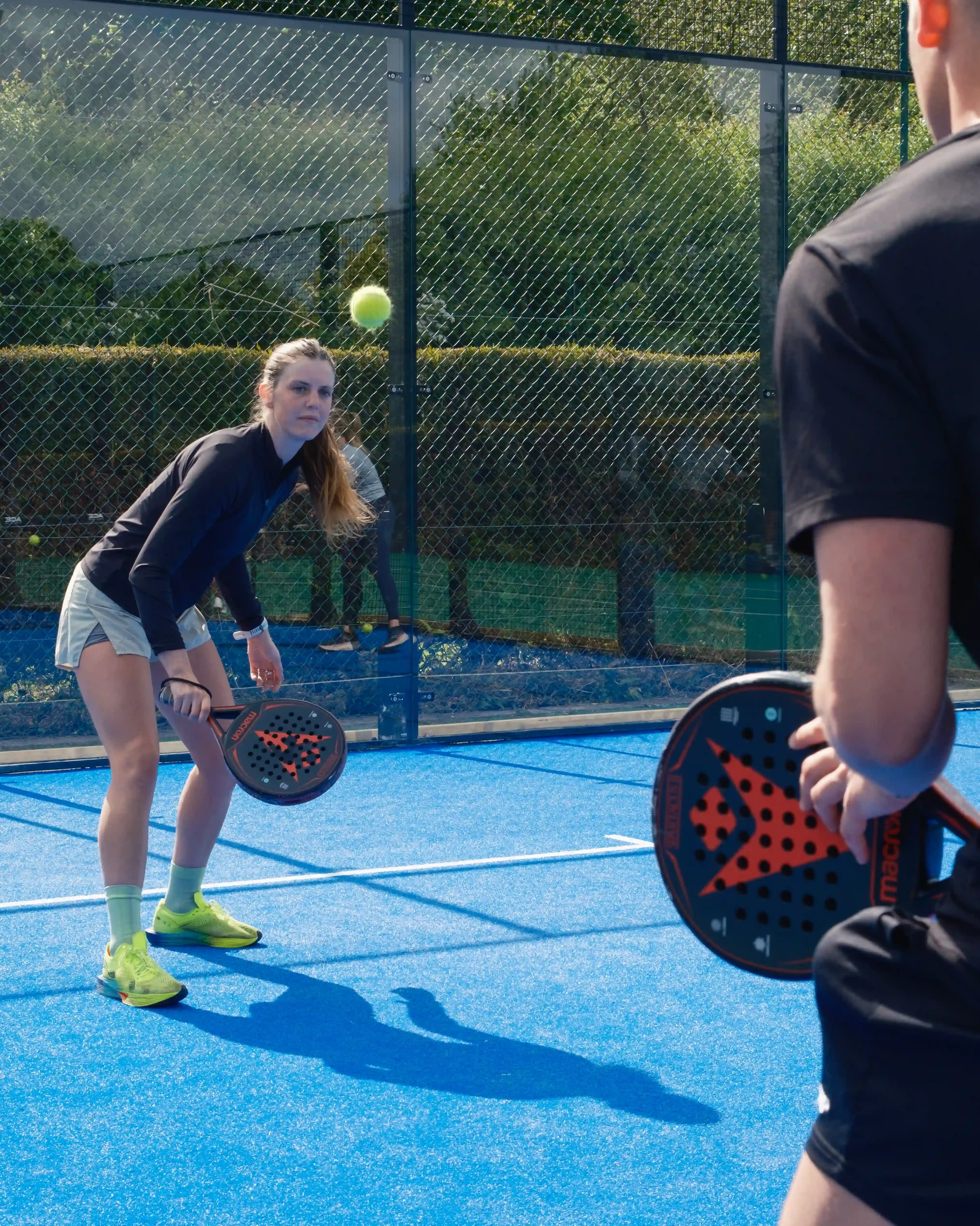 A dynamic, over-the-shoulder action shot on a vibrant blue padel court. A female player with a ponytail stands in an athletic ready position, eyes focused on a yellow padel ball in mid-air. She holds a black Macron racket with red geometric detailing