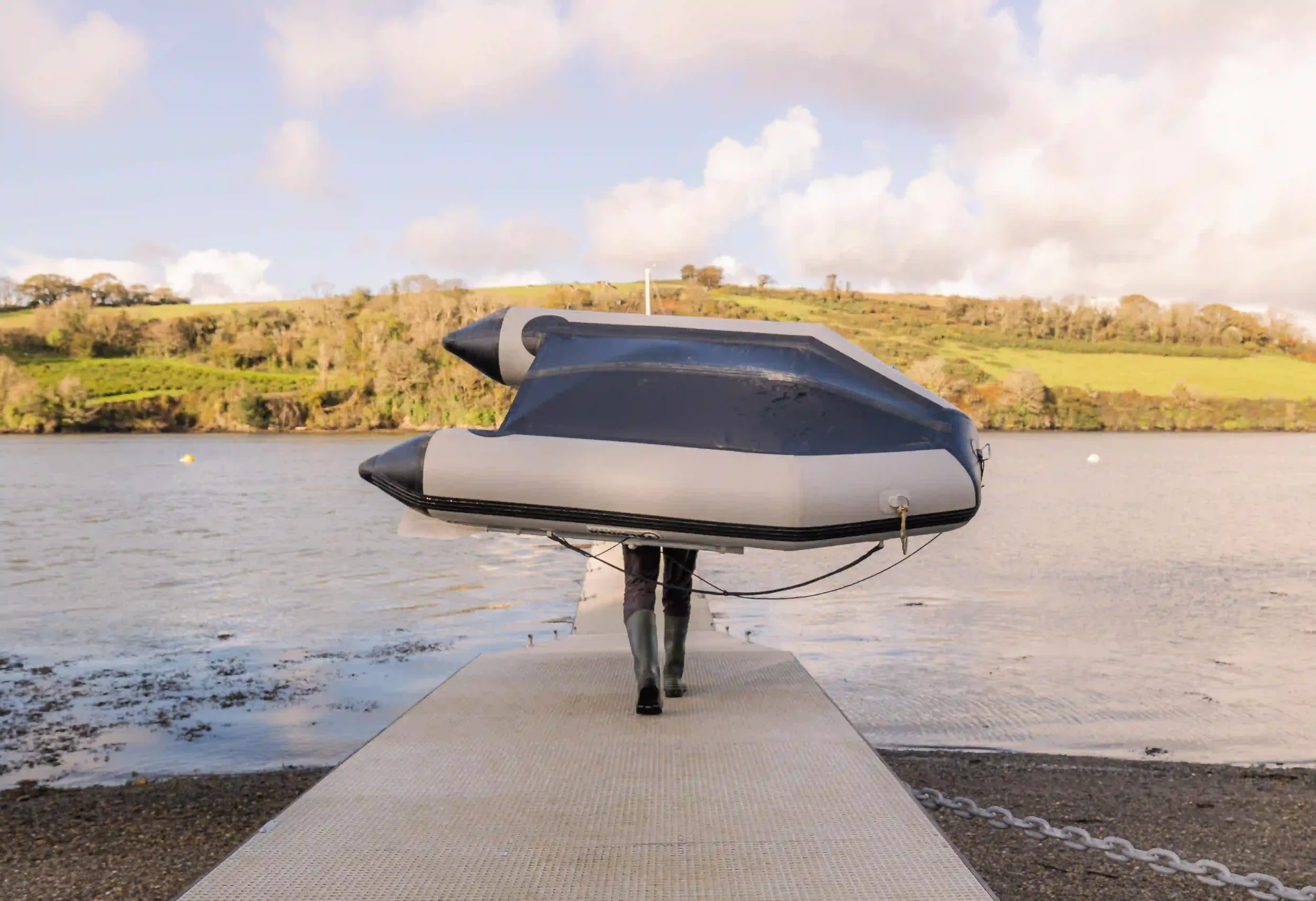 A person wearing green wellington boots walks away from the camera down a light-colored metal pier, carrying a large inflated black and grey boat over their head. The calm Fal River stretches out before them toward green, wooded hills under a bright,