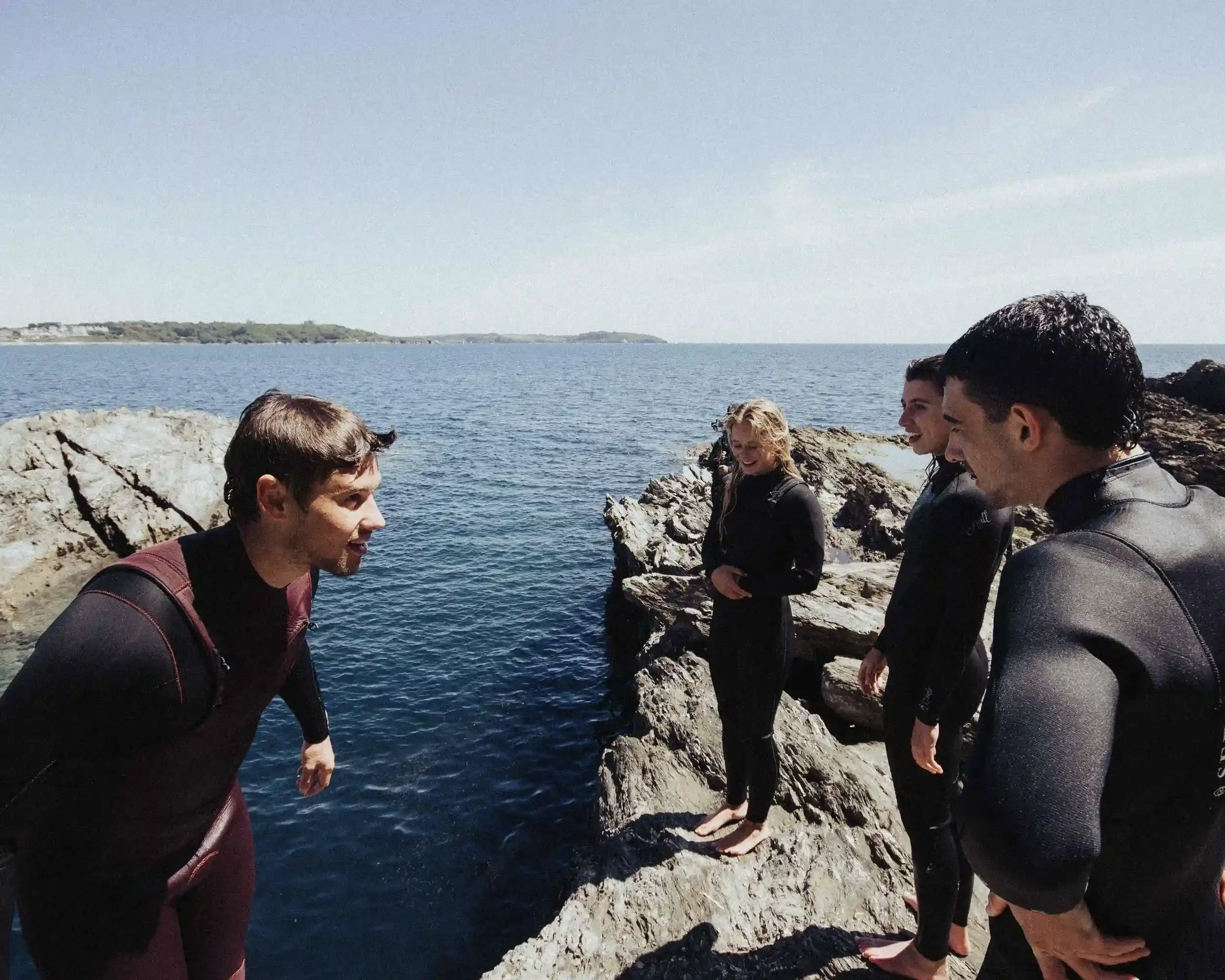 A candid photograph of four people, including singer-songwriter Ned Holland, standing on the edge of a rugged grey cliff in black and maroon wetsuits. They are looking at each other and smiling, preparing to jump into the clear blue ocean below. The 