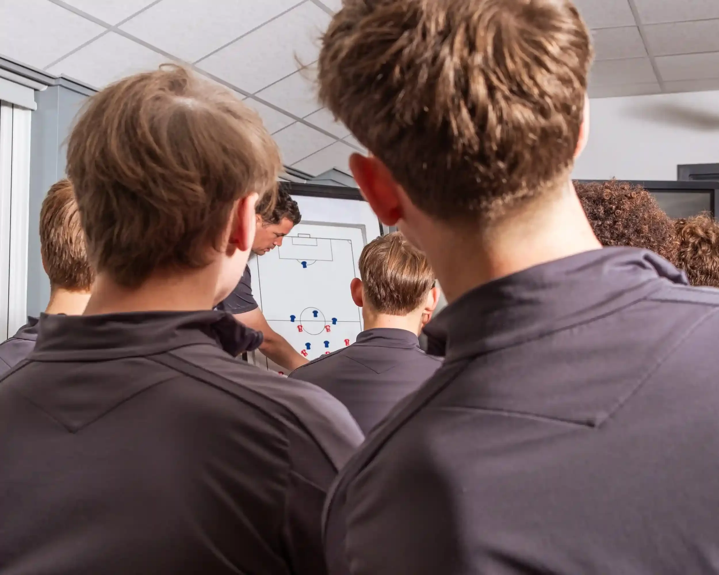 An over-the-shoulder shot of Wrexham AFC youth players wearing grey training kits, looking toward a tactical whiteboard. A coach is visible in the background, pointing to red and blue custom tactical magnets from Your Game Your Name arranged on a pit
