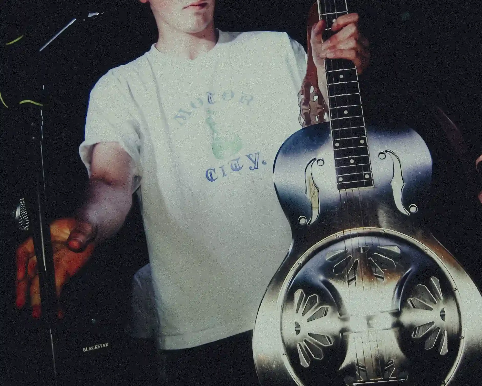 A gritty, high-contrast close-up of a metallic resonator guitar held by Ned Holland. The image focuses on the ornate metal body and f-holes of the instrument, with the musician's hand visible on the fretboard. He is wearing a white 'Motor City' graph