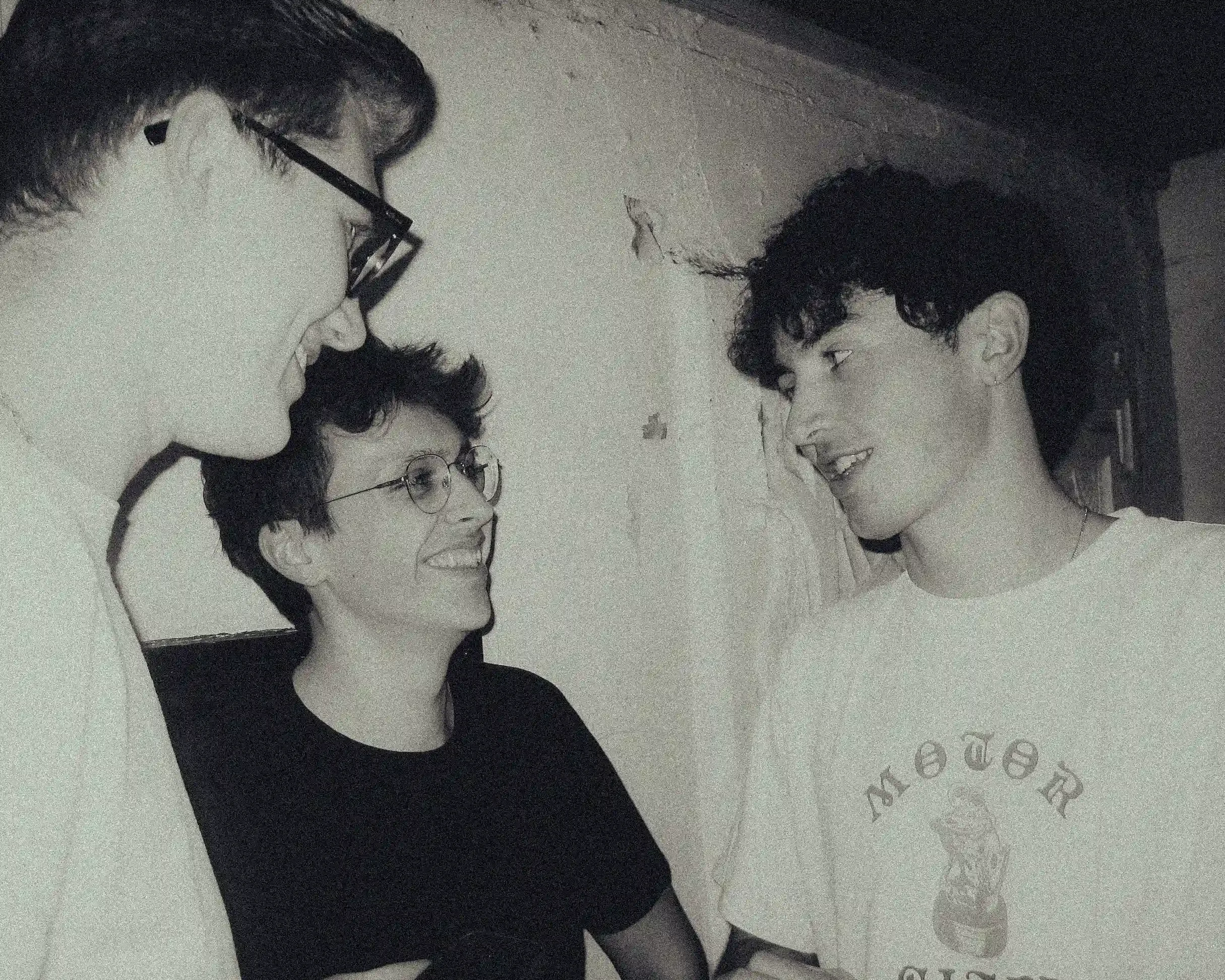 A grainy, black-and-white candid photograph of three young men backstage at a music venue. Ned Holland, on the right in a white 'Motor City' t-shirt, is looking toward his two bandmates and smiling. The two men on the left, one wearing glasses, are l