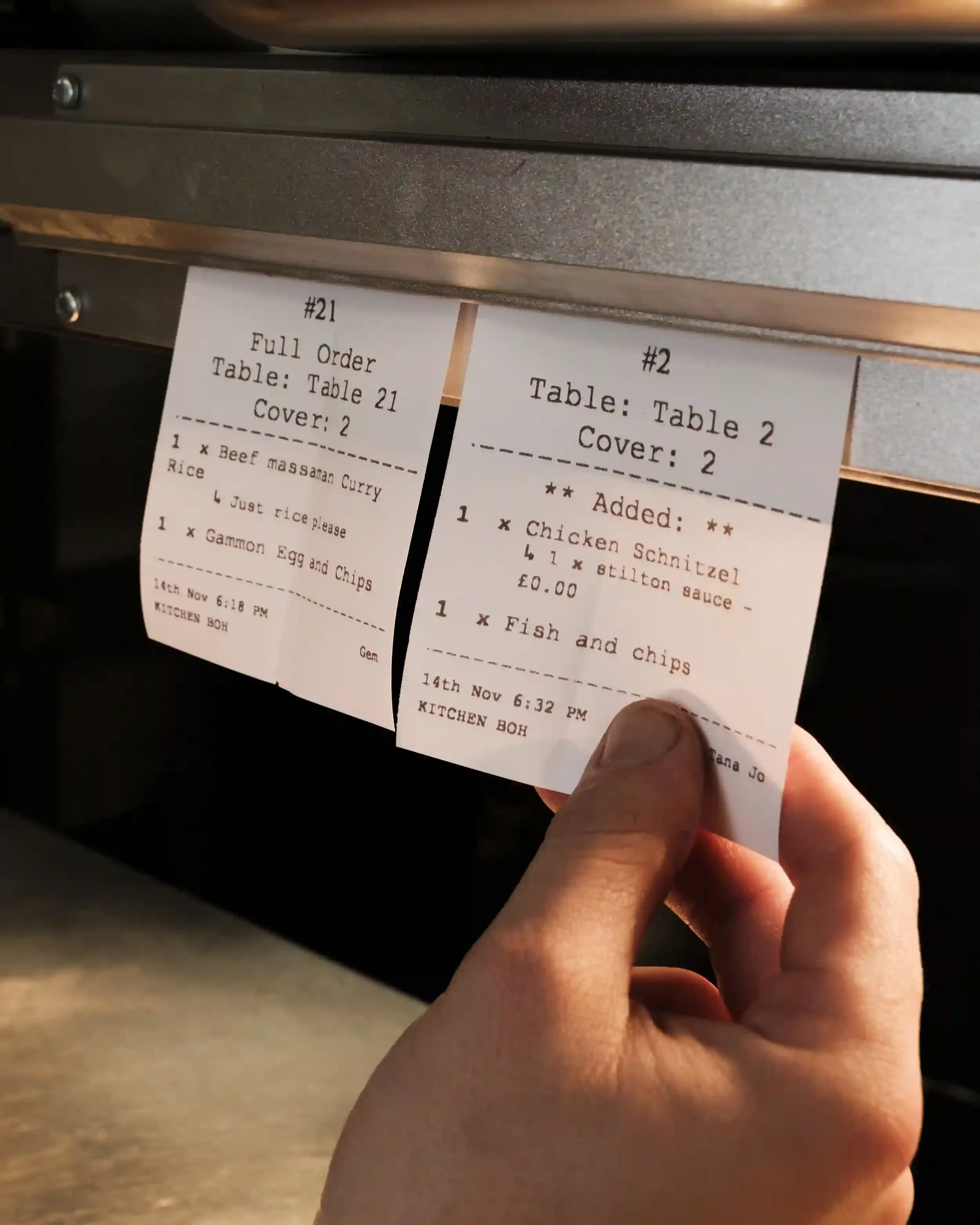 A close-up photograph of two white paper kitchen order tickets hanging from a metal rail at The Crown Inn, Lea. A hand is visible reaching for ticket #2, which lists 'Chicken Schnitzel' and 'Fish and chips.' Ticket #21 shows orders for 'Beef massaman