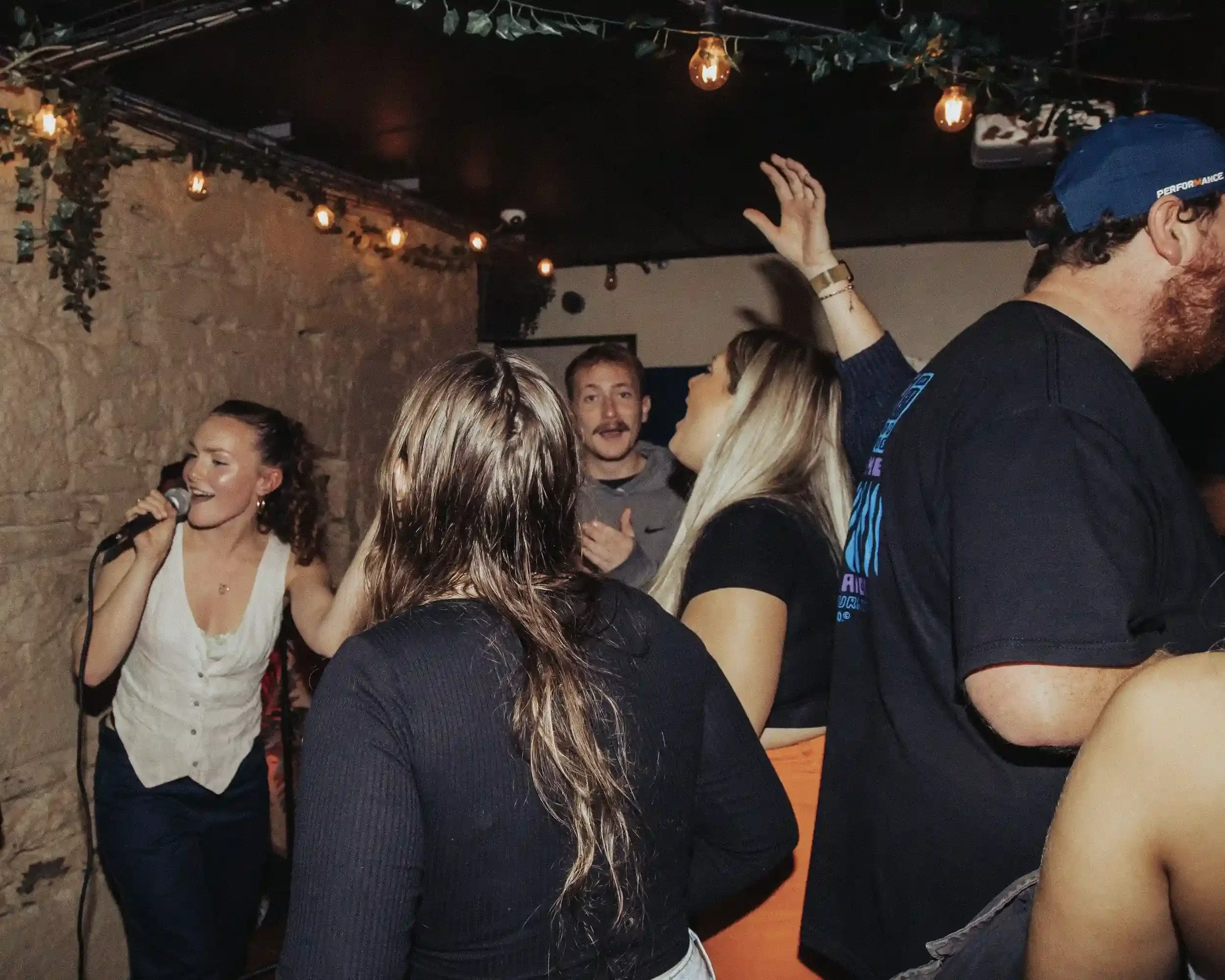 Authentic live music photography by Matthew Morgan at Small Ships bar. The image captures a female singer performing for a crowded room during a charity fundraiser. High-contrast flash emphasizes the raw, energetic '5-star dive bar' atmosphere and th