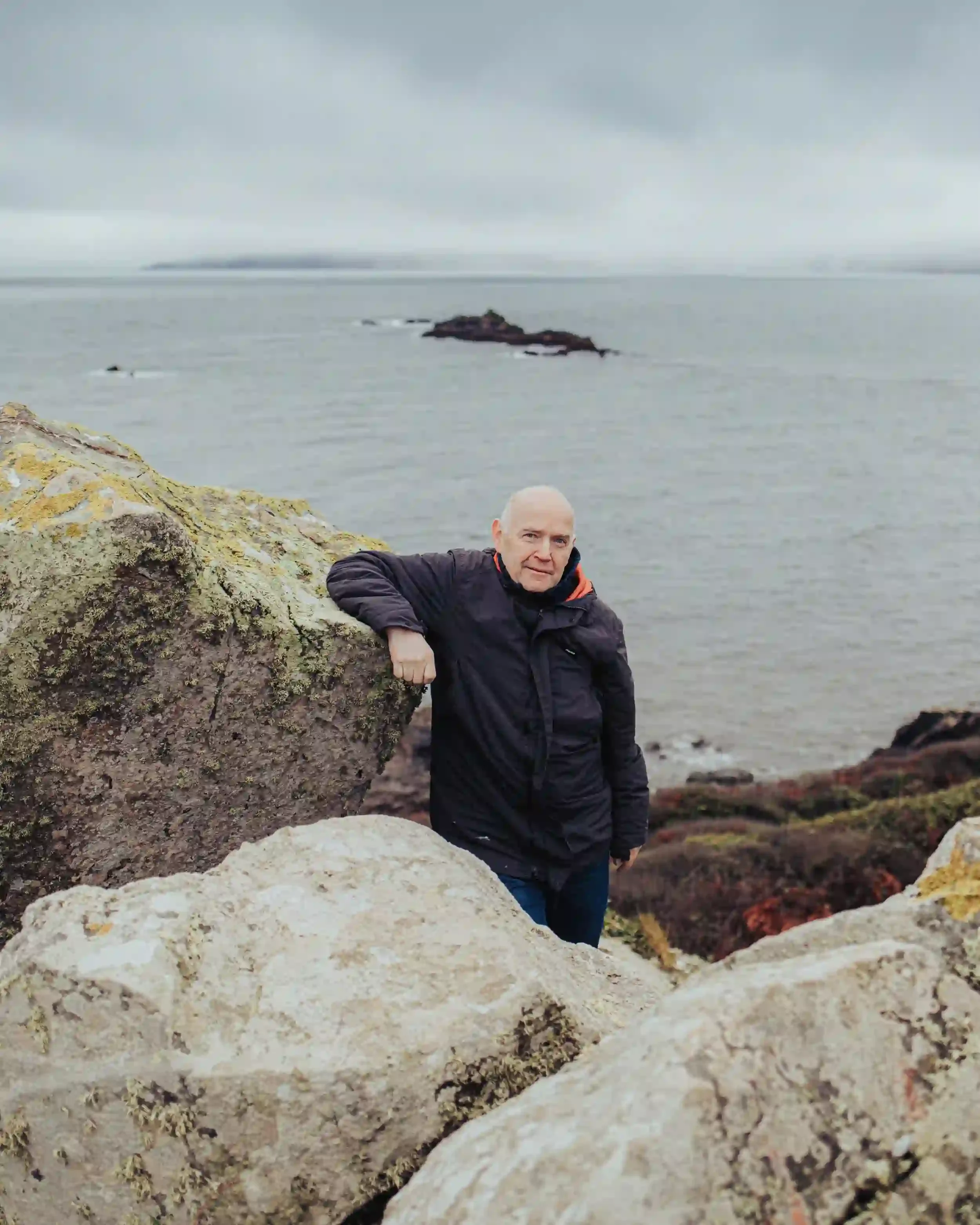A professional environmental portrait of artist David Mankin standing on a rocky cliffside in Cornwall. He is wearing a dark winter coat and leaning casually against a large, lichen-covered granite boulder. In the background, a grey, misty sea stretc