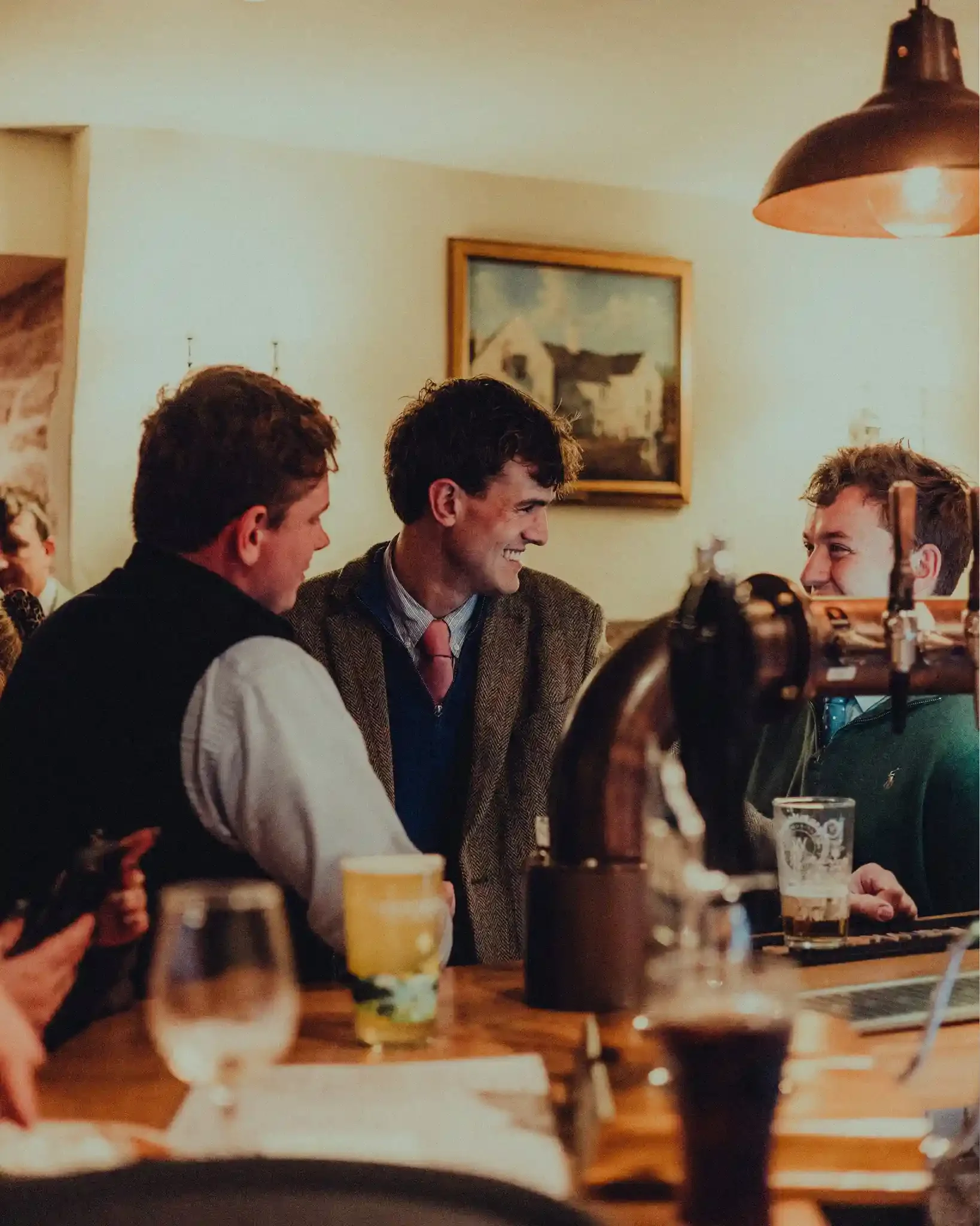 A warm and candid photograph of three men sharing a laugh at the bar of The Crown Inn in Lea. The central figure, wearing a tweed blazer over a blue sweater and pink tie, smiles broadly at a friend while holding a pint. The bar features traditional c