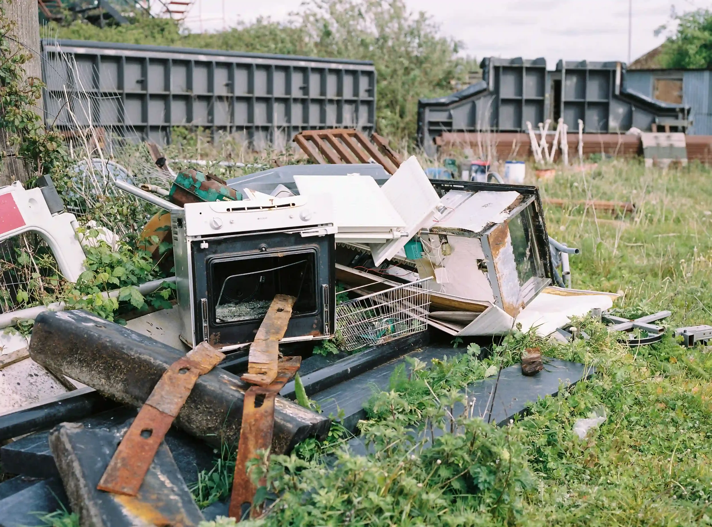 A pile of discarded household junk, including a broken white oven with its door open and rusted metal scraps, sitting in tall green grass in an industrial area of Sharpness, by Matthew Morgan.