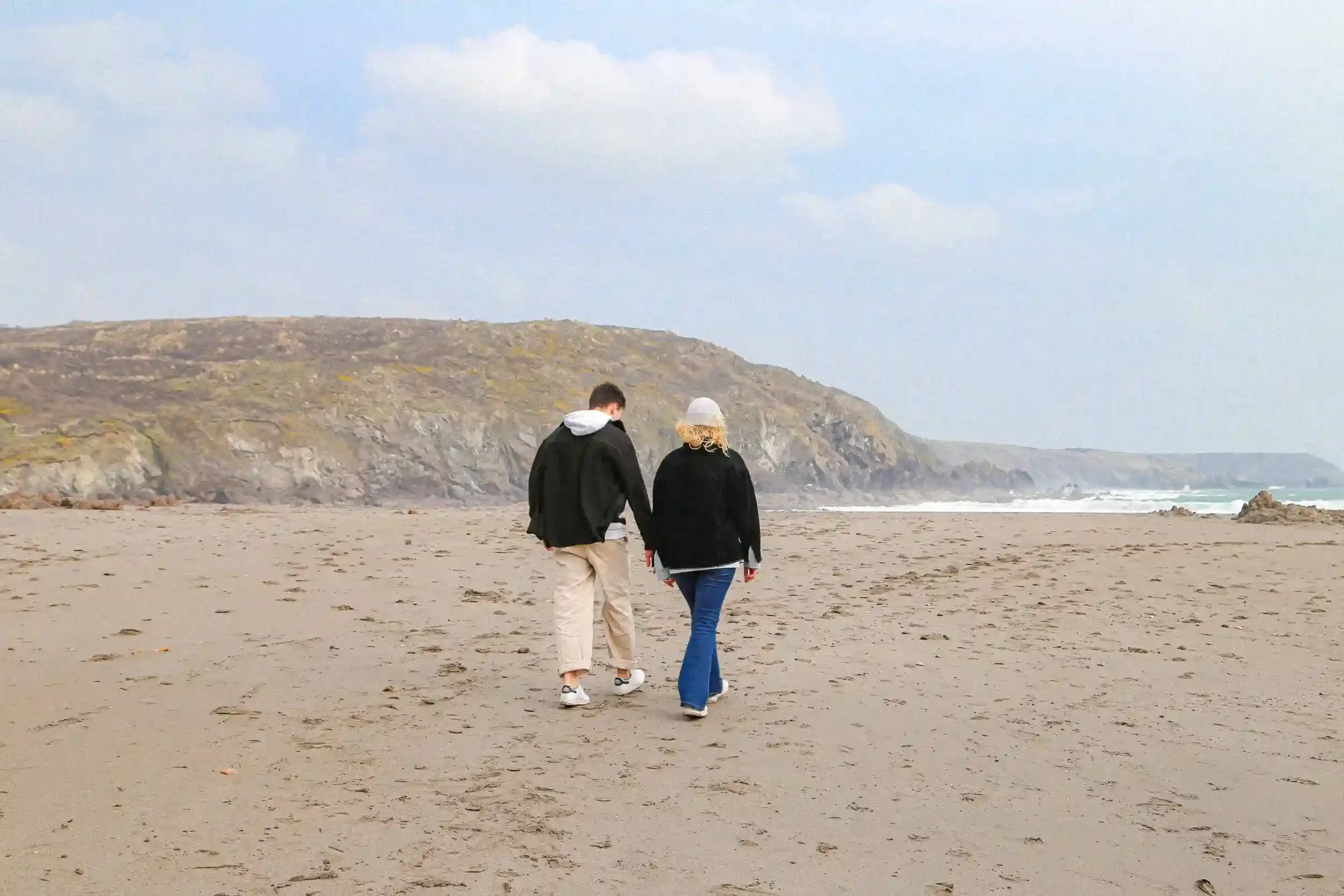 A wide-angle colour unit still from the film 'Lighter.' Two characters, seen from behind, walk across a wide, sandy beach in Cornwall. The man on the left wears a black jacket and cream trousers, while the woman on the right wears a black coat and bl