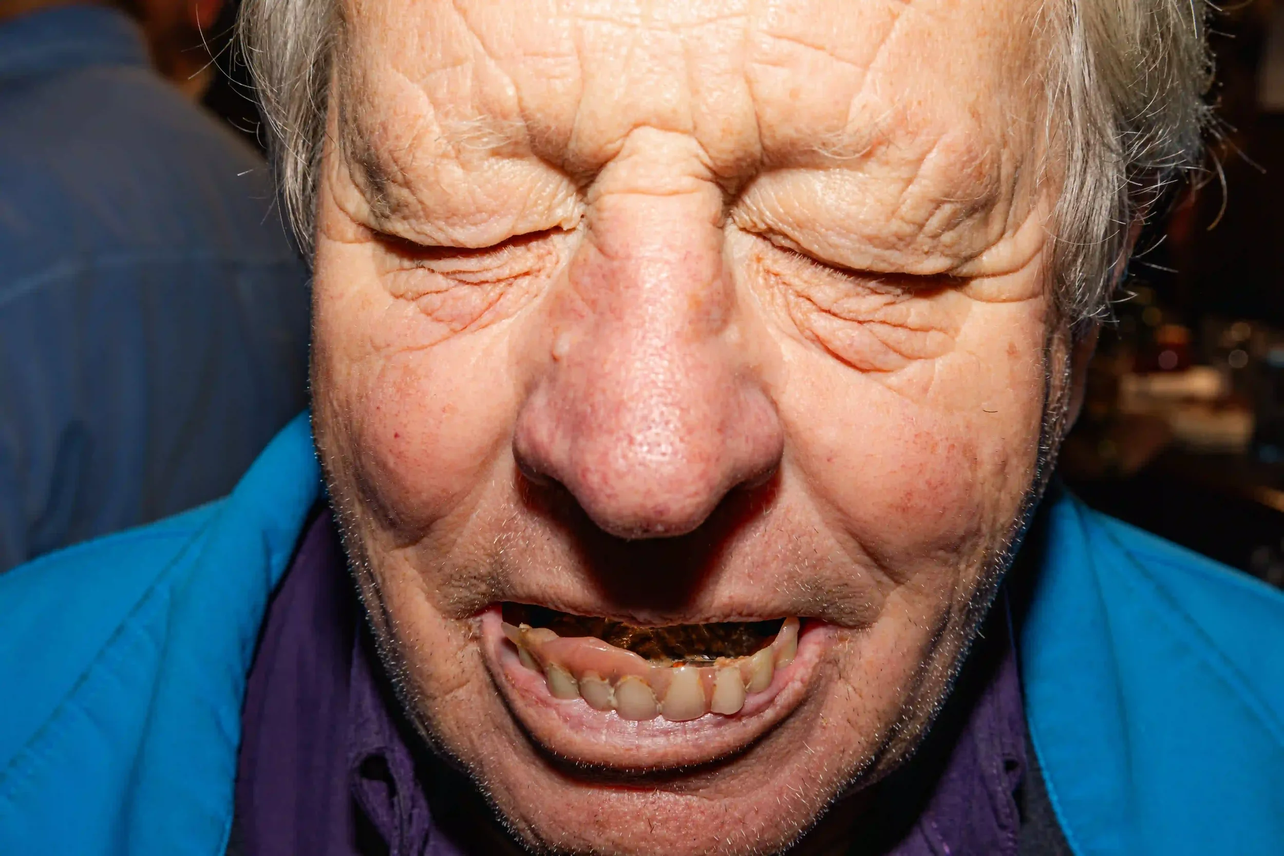 Extreme close-up of an elderly sailor's face at the Seaview Inn, Falmouth; his eyes are closed and he is laughing or grimacing to reveal a set of worn false teeth, with the bright blue fabric of his jacket visible in the frame, by Matthew Morgan.