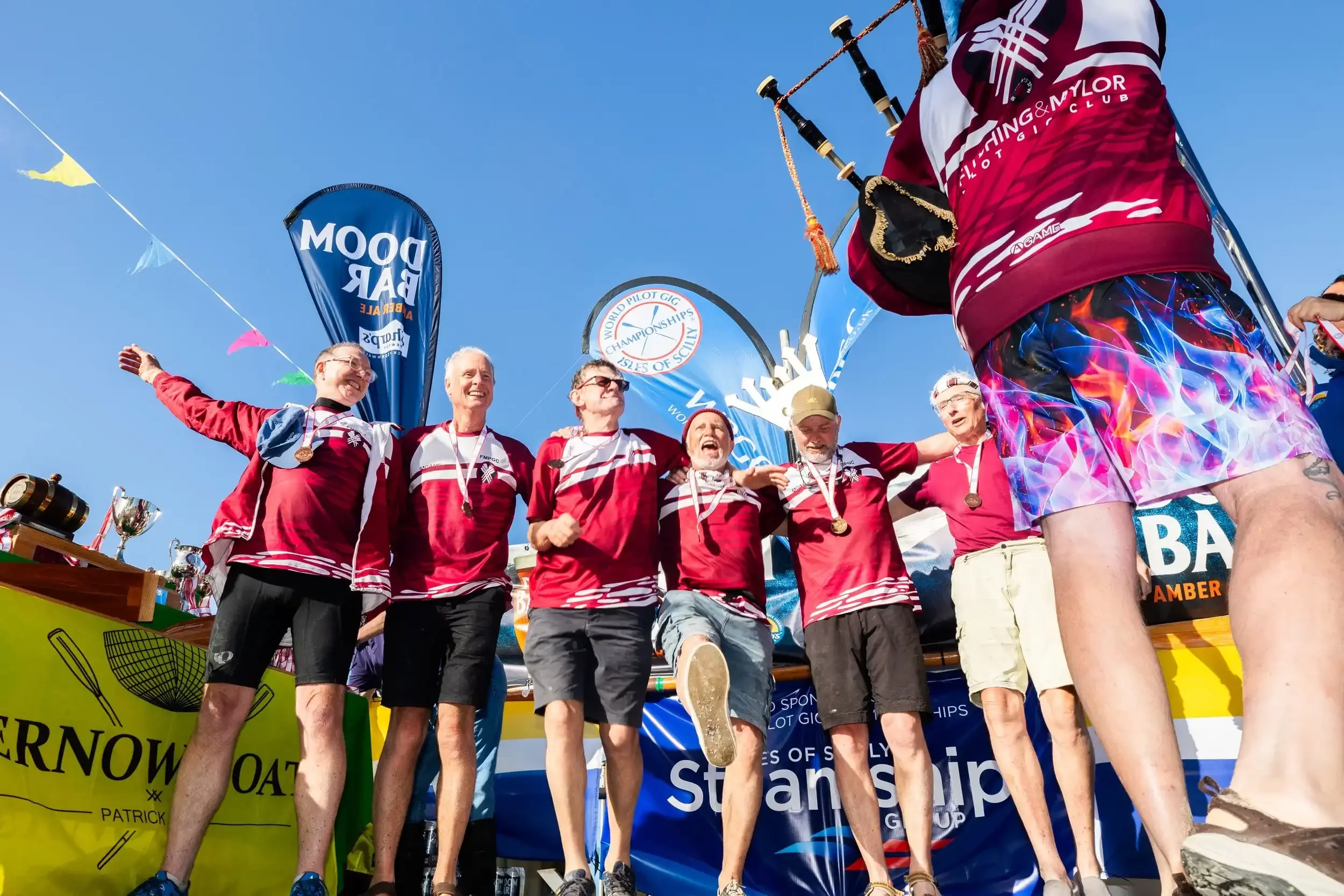A low-angle, wide shot of the Flushing & Mylor Pilot Gig Club crew celebrating on an outdoor podium. Five men in maroon and white team shirts stand with their arms around each other, wearing bronze medals and shouting in joy. To the right, the back o