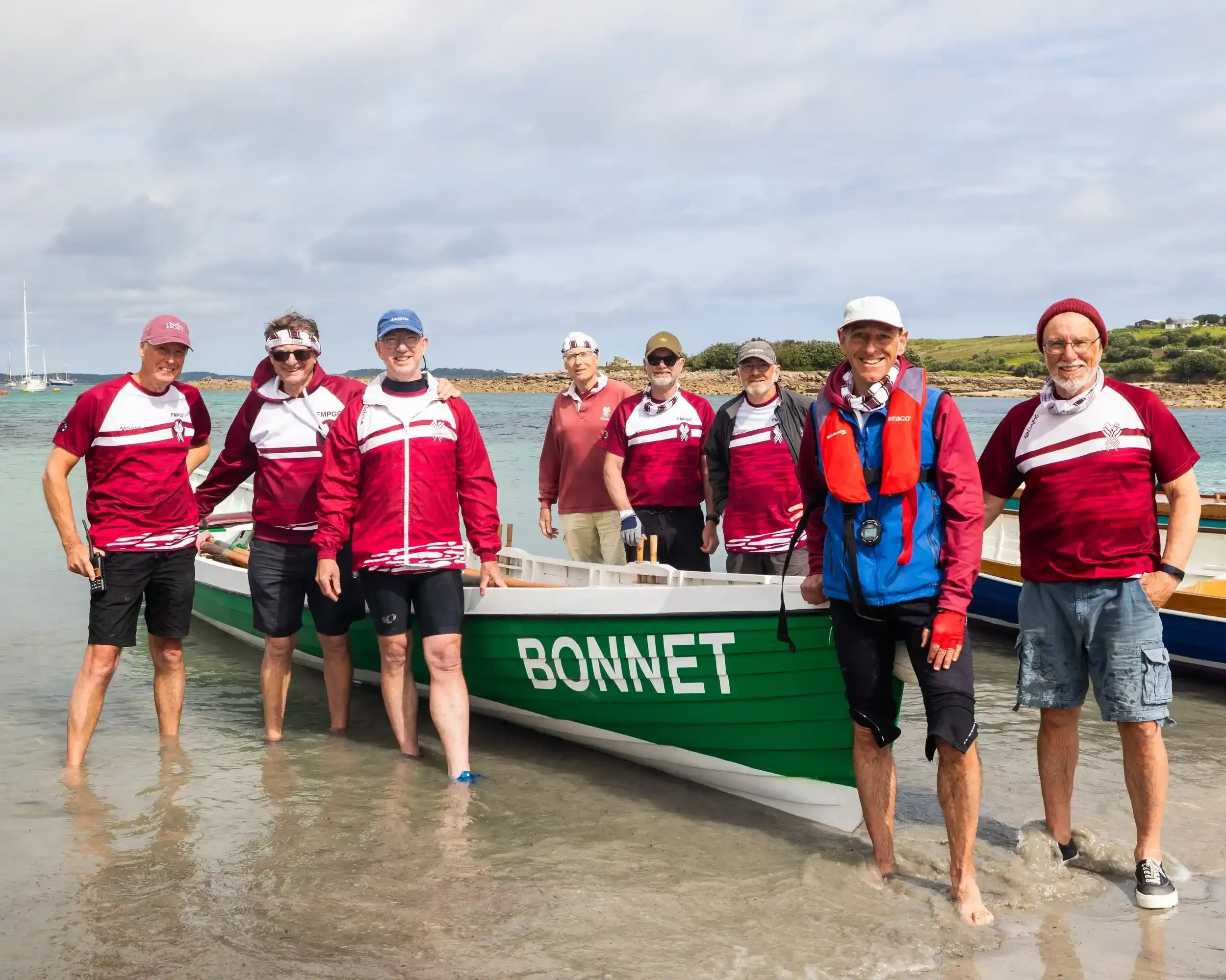 A group portrait of eight male rowers from the Flushing & Mylor Pilot Gig Club standing in knee-deep clear water next to their boat, the 'Bonnet'. The boat is bright green with white trim and the name 'BONNET' painted in bold white letters on the bow