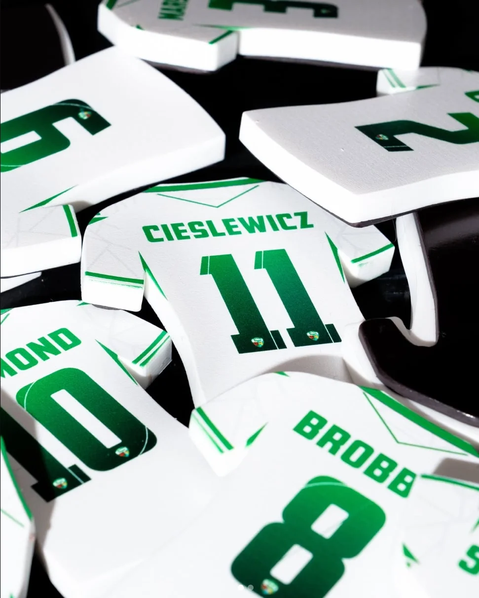 A bright, close-up photograph of several white jersey-shaped magnets piled together. The central magnet is in sharp focus, featuring the name 'CIESLEWICZ' and the number '11' in a green gradient. Other visible magnets show 'BROBBEL 8' and 'REDMOND 10