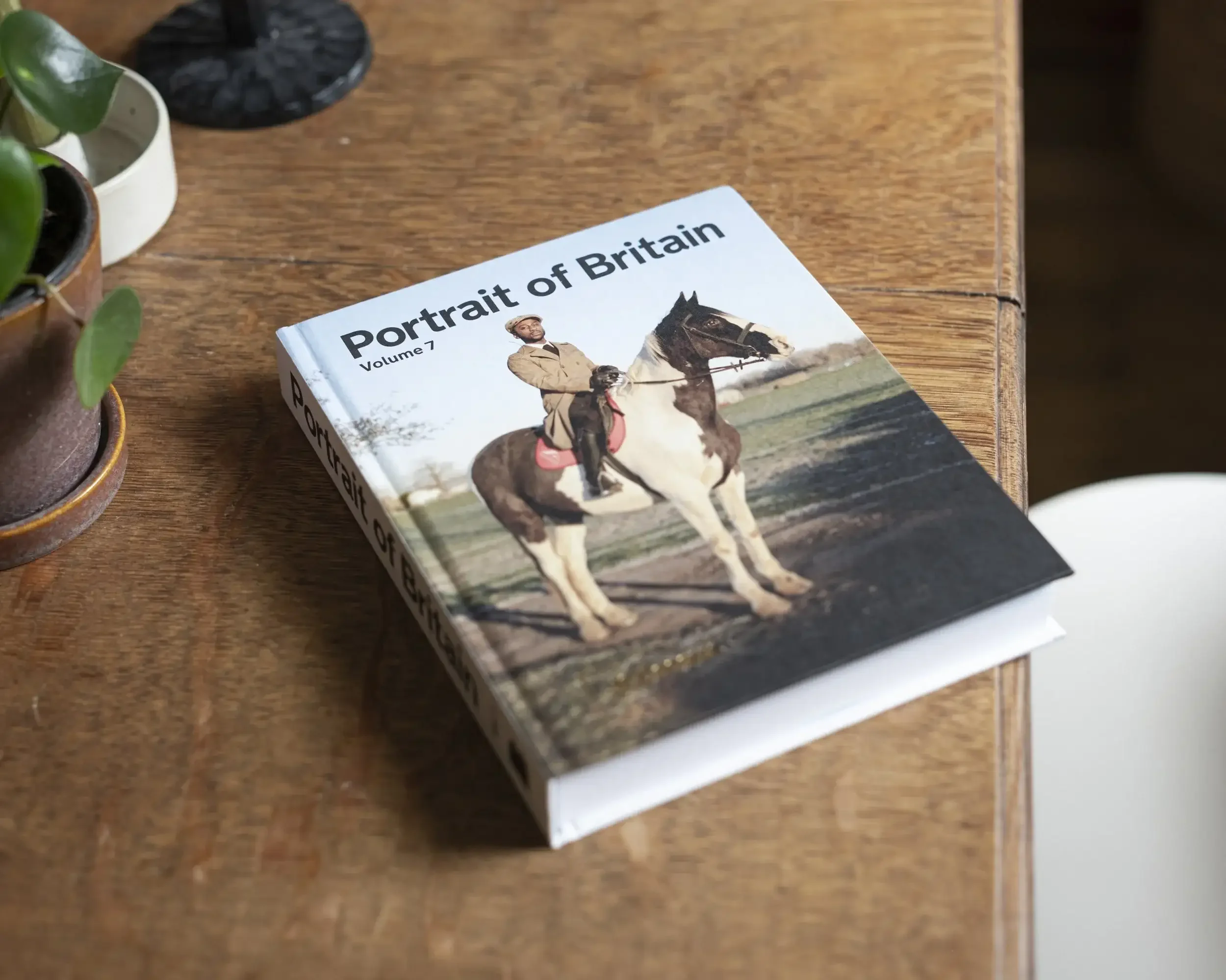 The front cover of the 'Portrait of Britain Volume 7' photobook resting on a wooden table. The cover features a photograph of a man in a brown suit sitting on a brown and white horse. The title 'Portrait of Britain Volume 7' is printed in bold black 