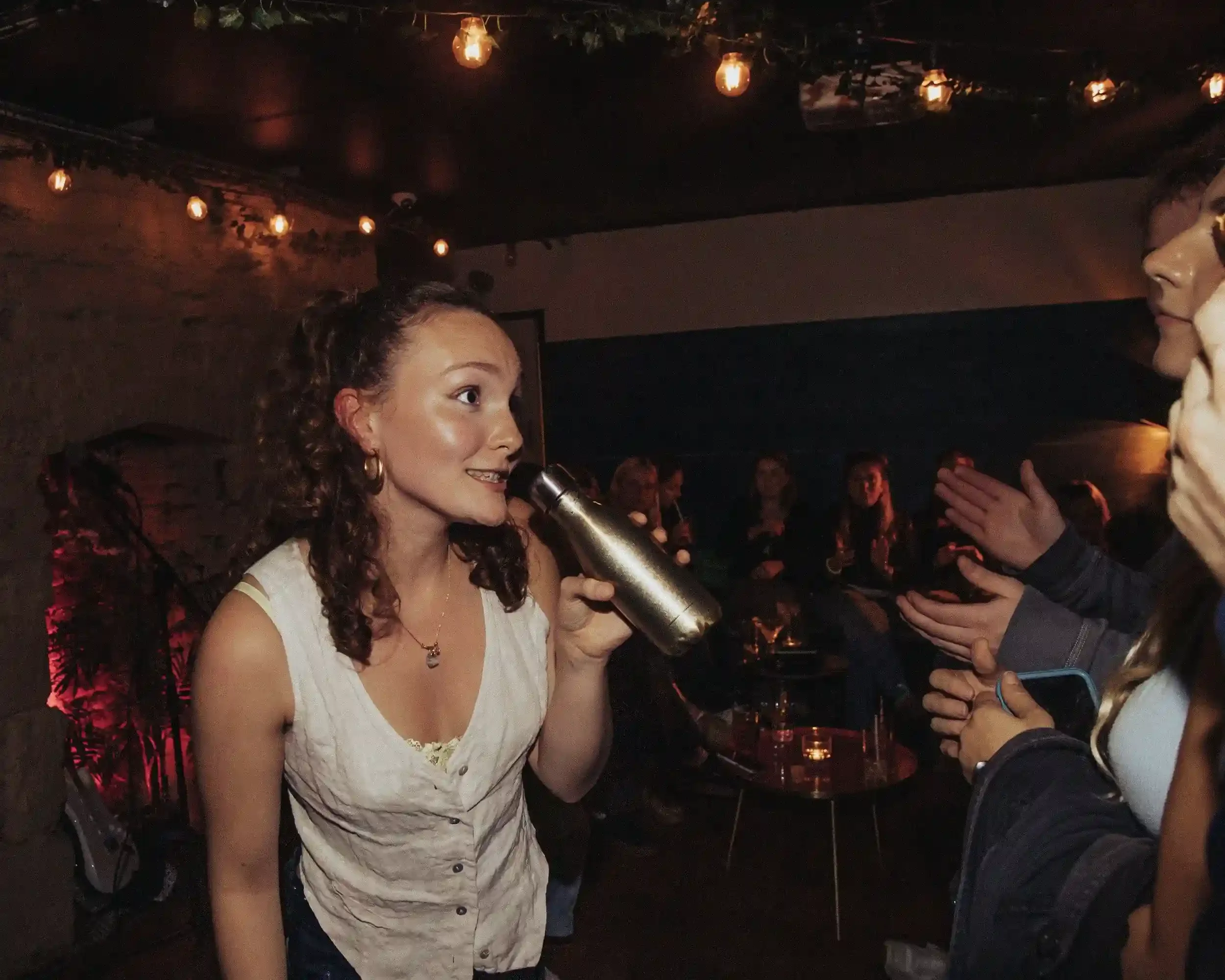 A candid, high-contrast shot of a woman in a white waistcoat holding a metallic gold water bottle while speaking to someone off-camera inside Small Ships bar. She is smiling and engaged, with a blurred audience and warm festoon lights in the backgrou