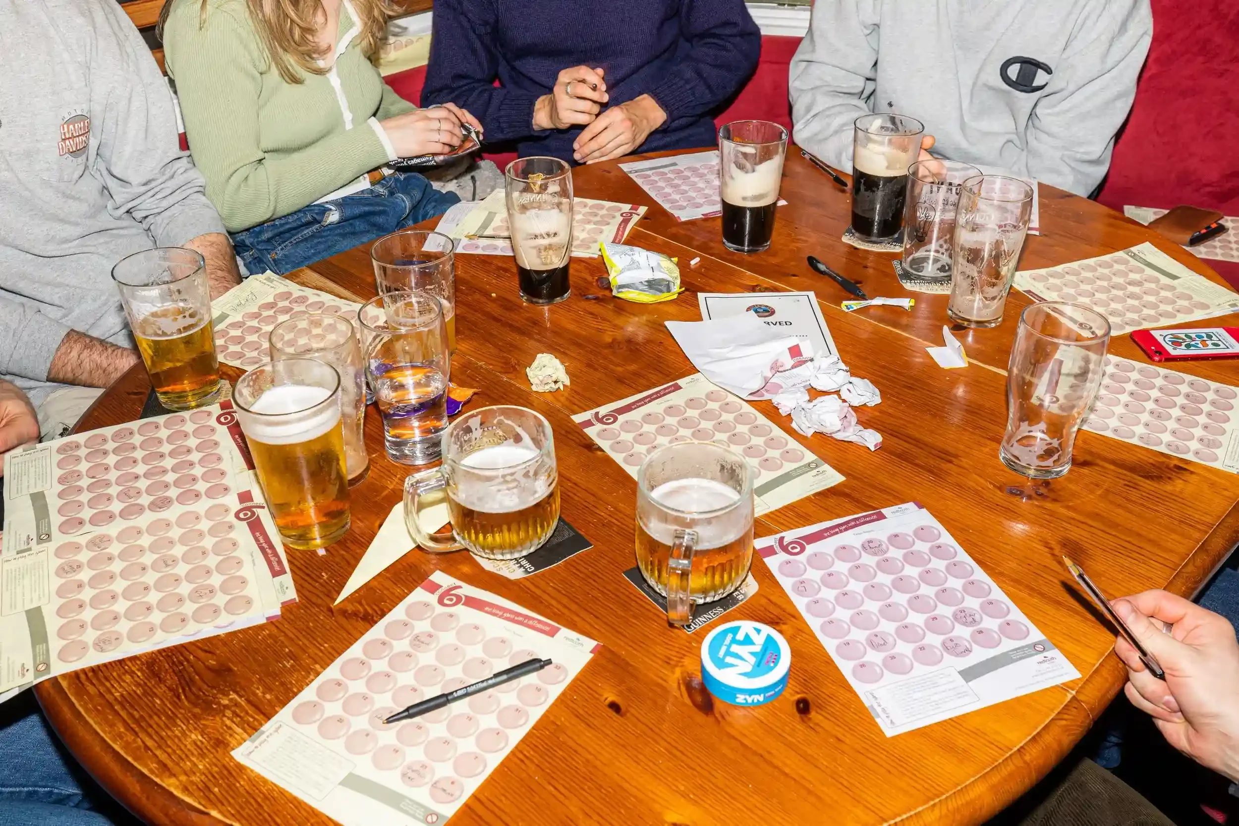 A busy wooden pub table covered in Quiz Go sheets and pints of beer during a community quiz night at the Seaview Inn, by Matthew Morgan.