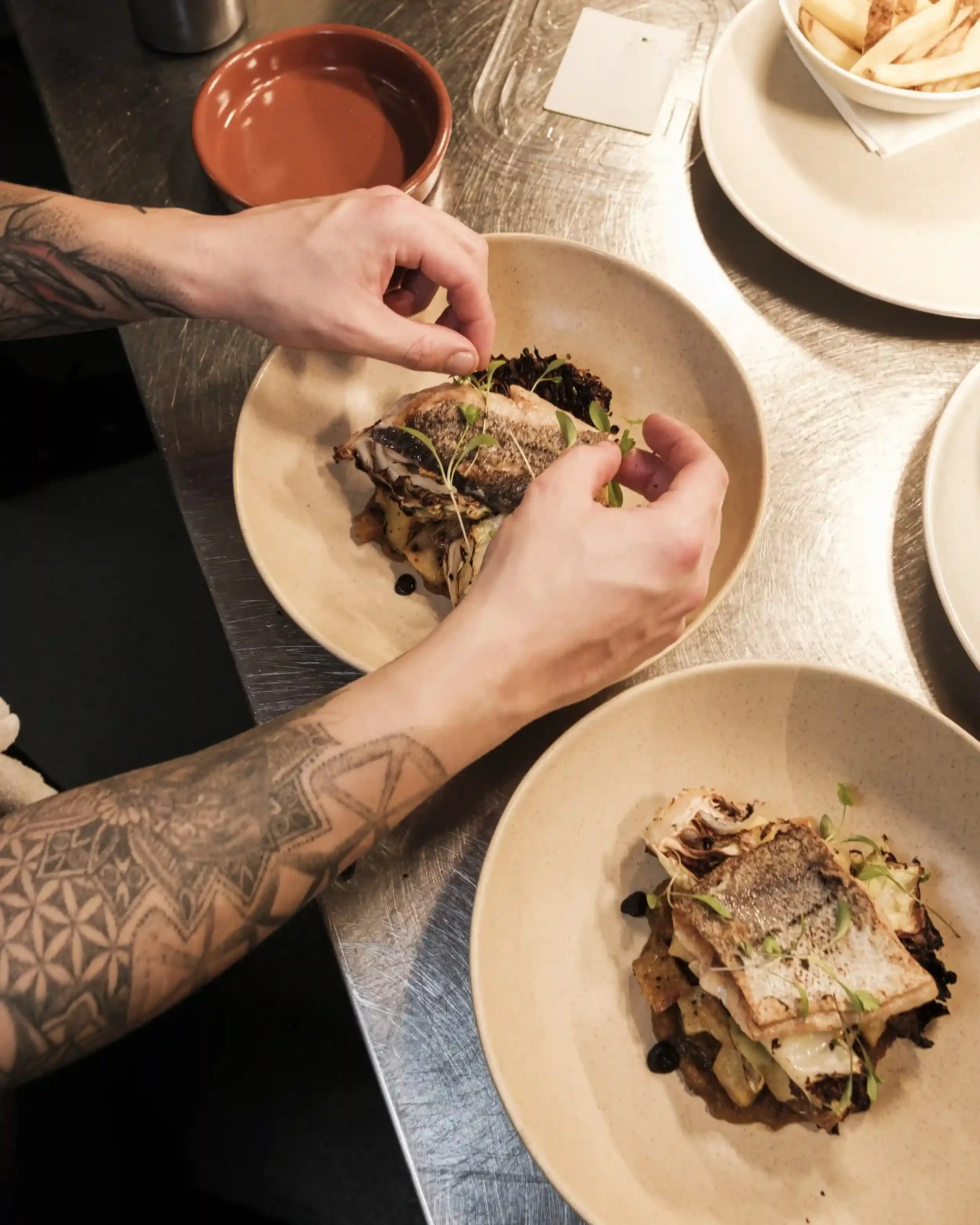 A top-down, close-up photograph of a chef’s hands garnishing a plate of pan-seared fish with delicate green micro-herbs. The chef, whose arms feature intricate geometric tattoos, is carefully placing the garnish on one of two identical plates. The di