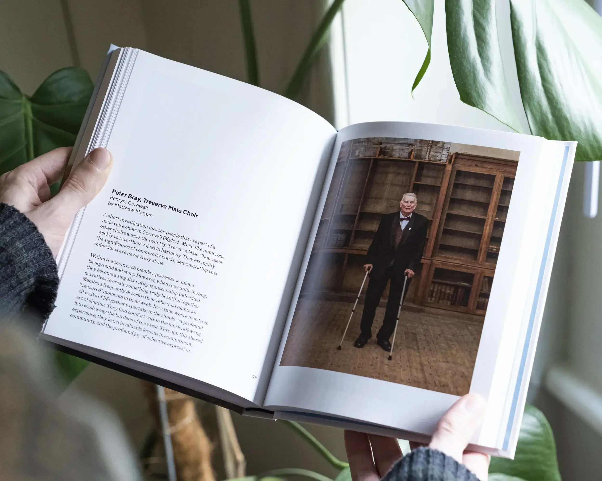A person's hands holding open the 'Portrait of Britain Vol. 7' photobook. The left page features text titled 'Peter Bray, Treverva Male Choir' by Matthew Morgan, describing the community significance of Cornish male voice choirs. The right page shows