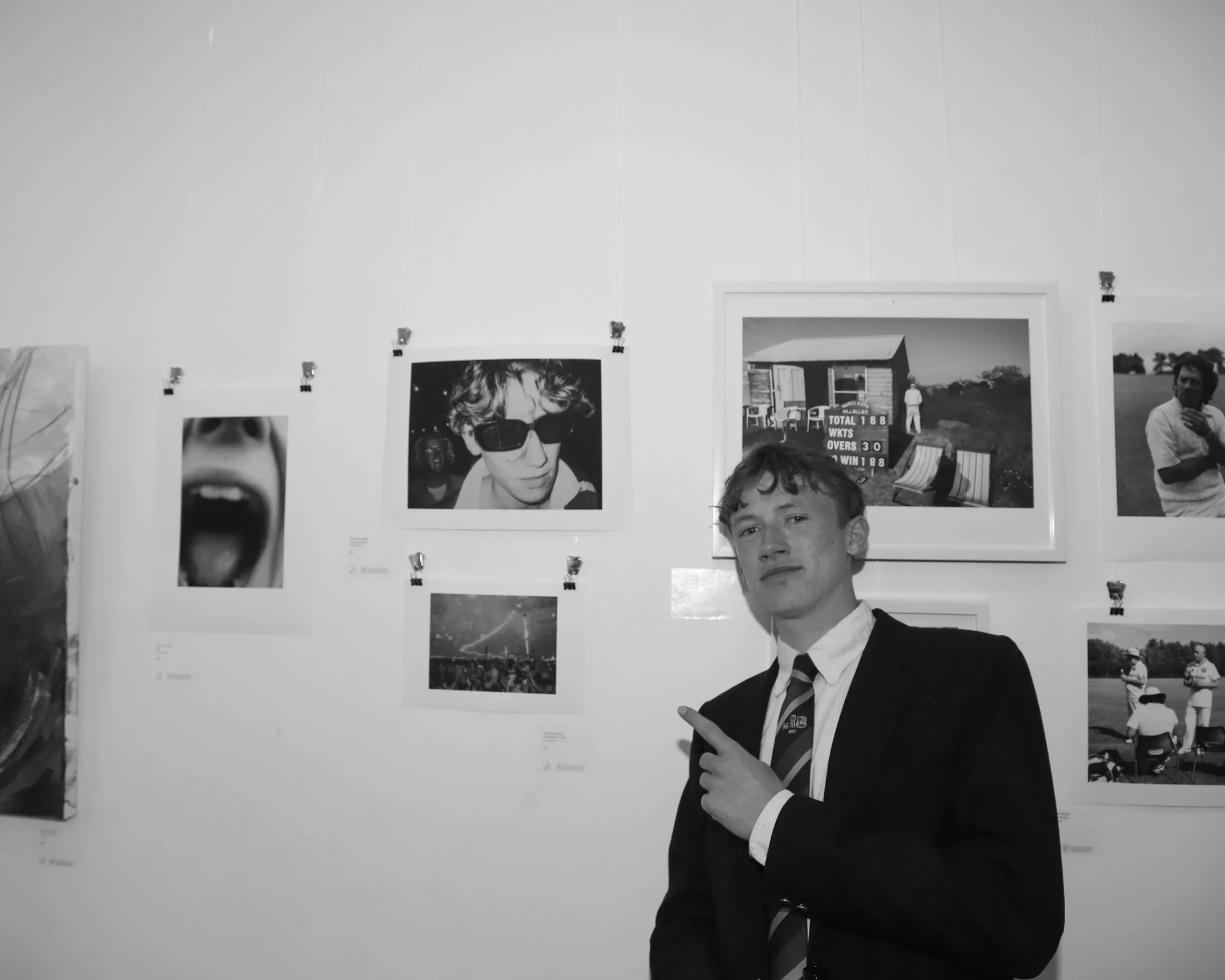 A black-and-white photograph of a young man named Henry Jones at the '2021-2024' exhibition at The Poly, Falmouth. He is wearing a dark suit and a striped tie, pointing with his right hand toward a framed photograph of himself on the gallery wall. Th