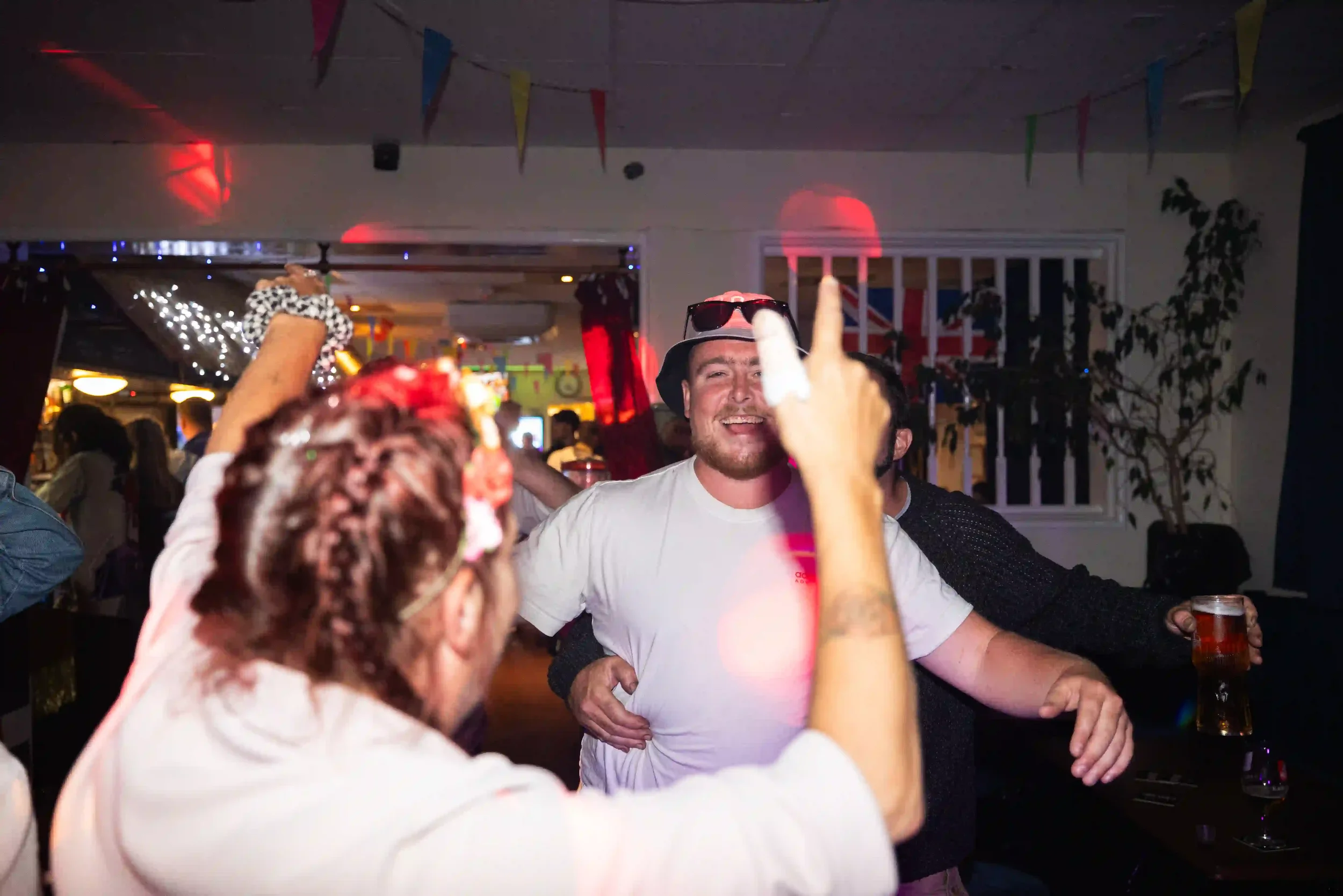 A festive interior scene at The Arwenack Club in Falmouth; a man in a bucket hat and sunglasses smiles while dancing, surrounded by others with their arms raised. Colorful bunting hangs from the ceiling, and red disco lights flare across the room, by