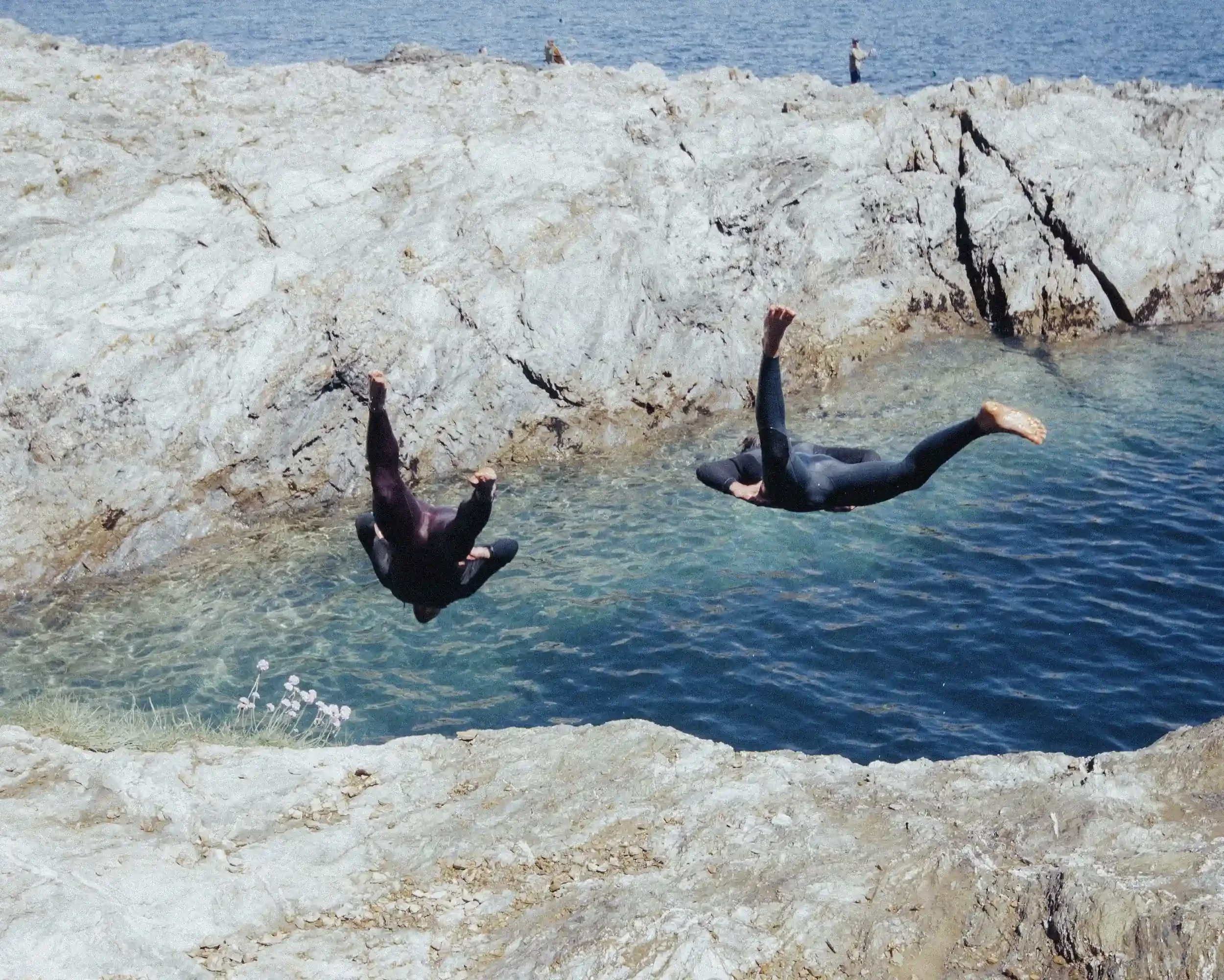 A high-action photograph of two people in black wetsuits performing backflips off a light grey rocky cliff into a bright blue ocean cove. The water is clear and turquoise near the shore, with pink flowers visible in the bottom left corner. This actio