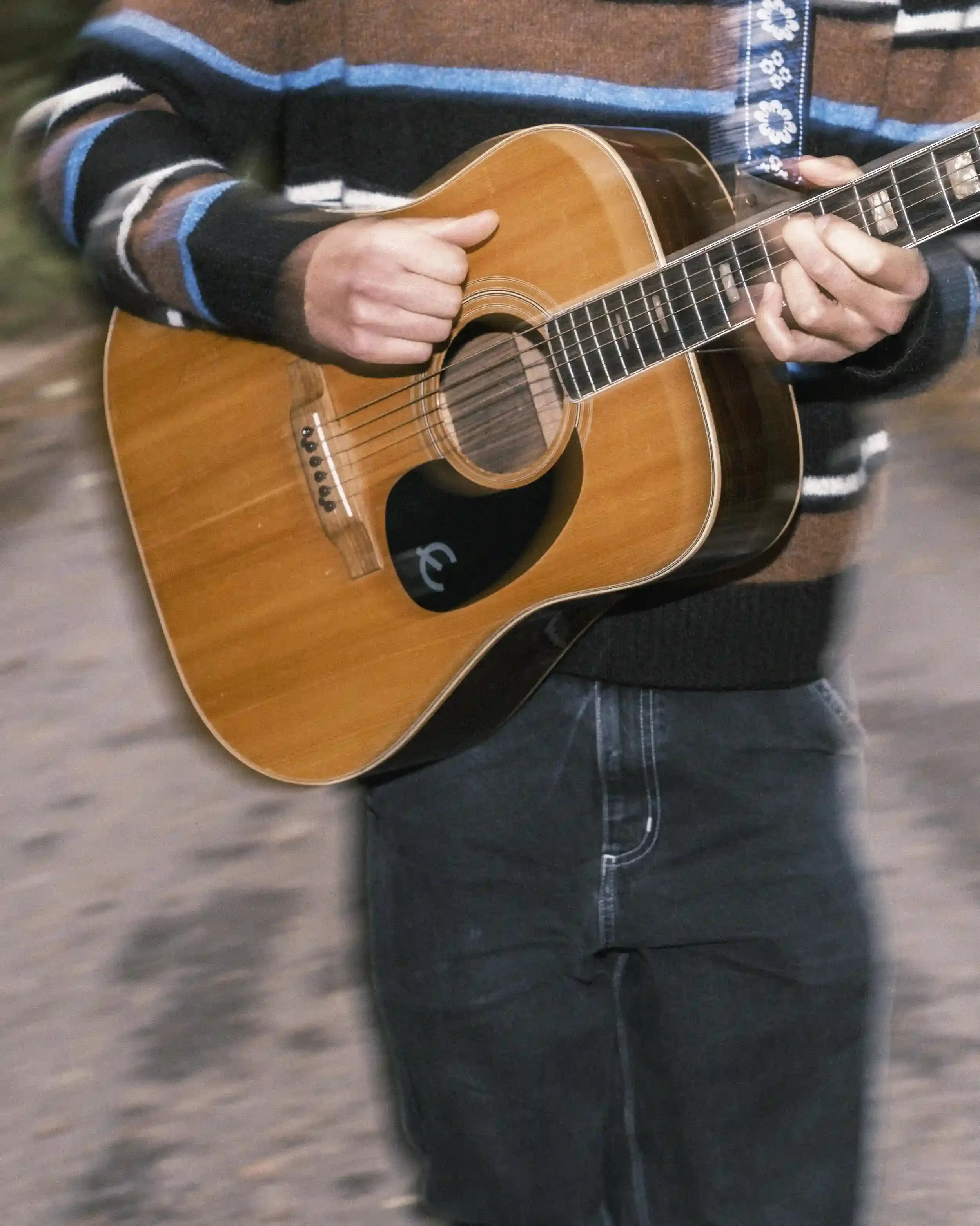 A close-up, slightly blurred action shot of musician Ned Holland playing an acoustic guitar. The focus is on the light-wood body of the guitar, featuring a black pickguard with a white 'E' logo. Holland's left hand is visible on the fretboard, while 