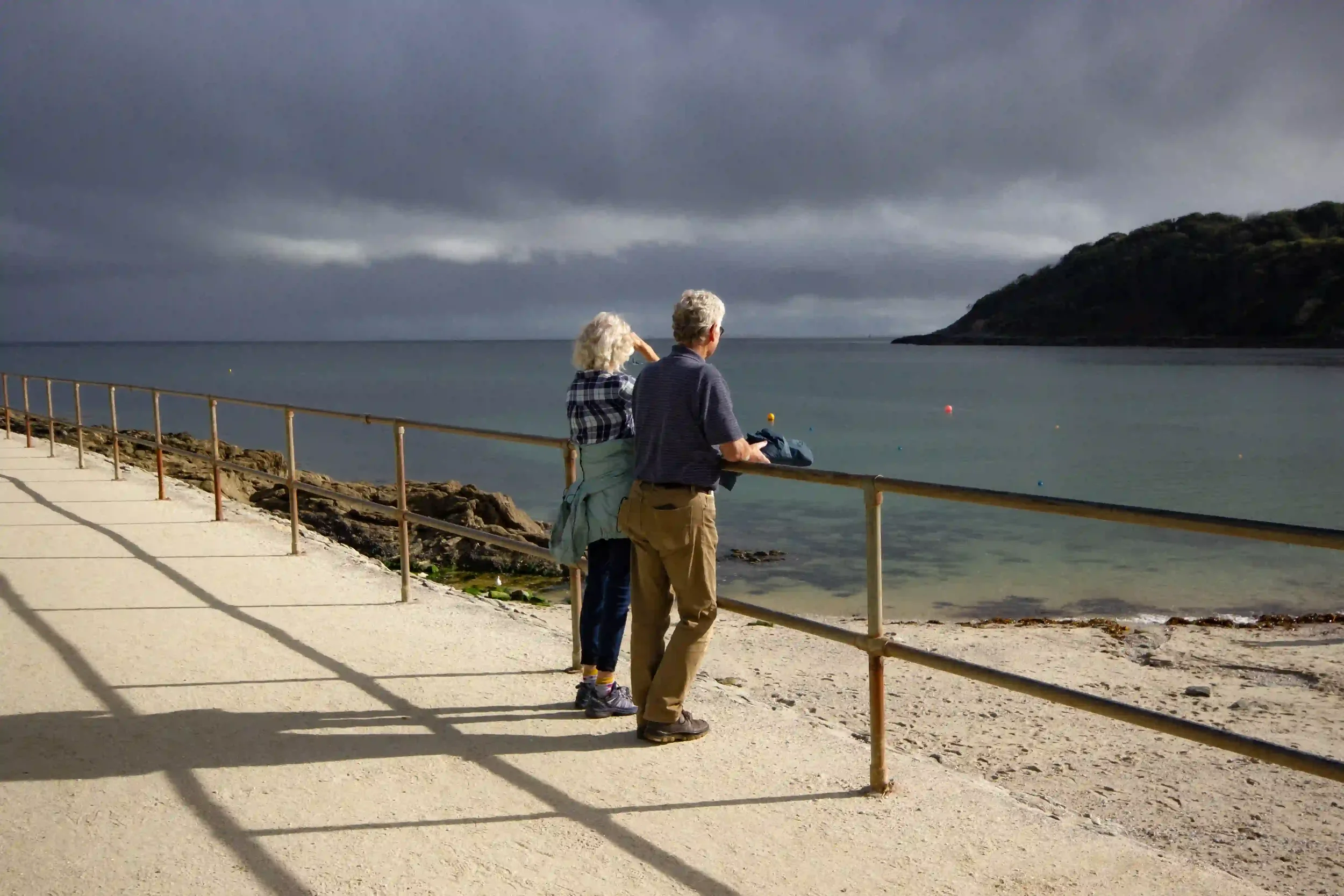 An older couple leaning against a rusted metal railing on a sunlit concrete path, looking out over the turquoise waters of Swanpool Bay in Falmouth, Cornwall. A dark, moody rain cloud hangs over the green headland in the distance, by Matthew Morgan.