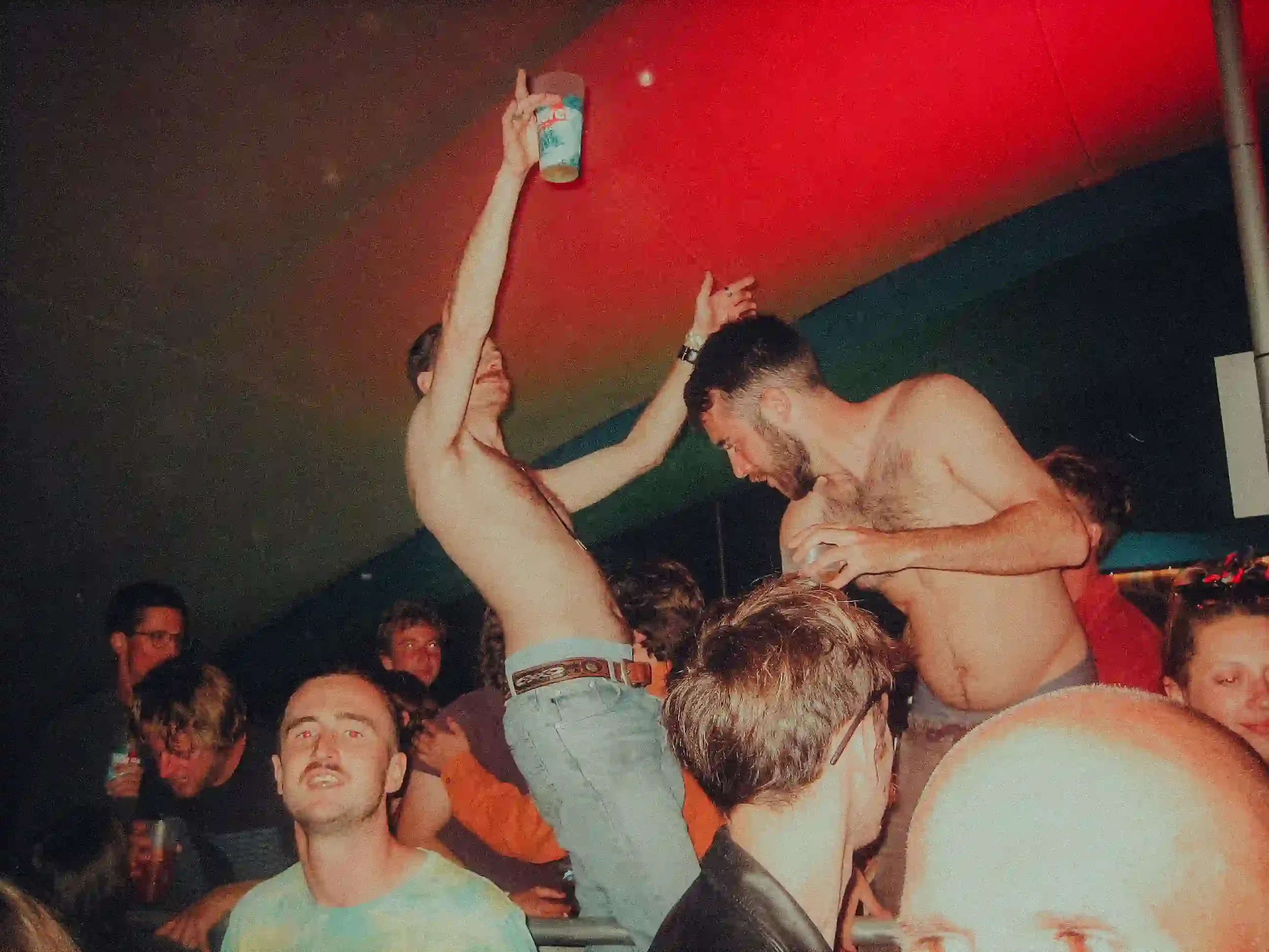 A candid, high-energy shot inside SkyBar, St Agnes, under a saturated red and green ceiling. Two shirtless men are dancing in the crowd, one with his arm raised holding a blue plastic cup, while other festival-goers look on in a dimly lit, crowded at