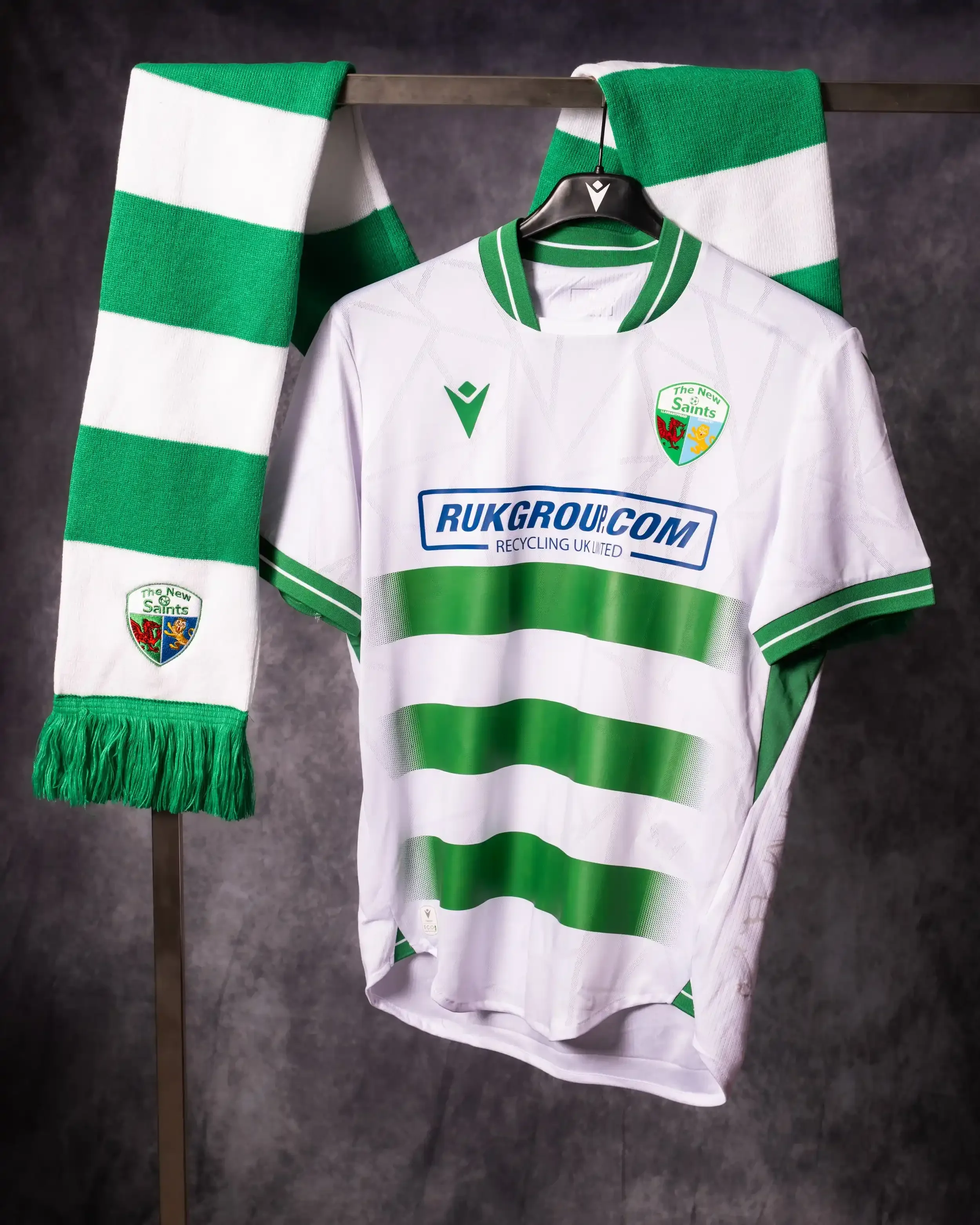 A professional studio product shot of The New Saints FC (TNS) home kit. A white jersey with green horizontal hoops and a subtle geometric pattern is displayed on a metal rack. A matching green and white striped bar scarf with the TNS crest hangs besi