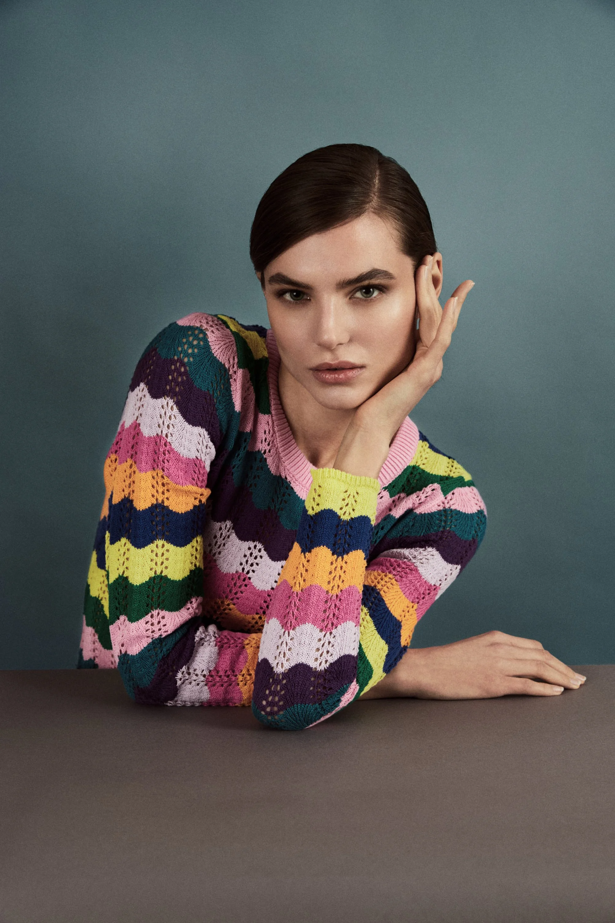 A woman with short, dark hair wearing a colorful, striped, crochet sweater resting her chin on her hand, seated at a table against a plain, teal background.