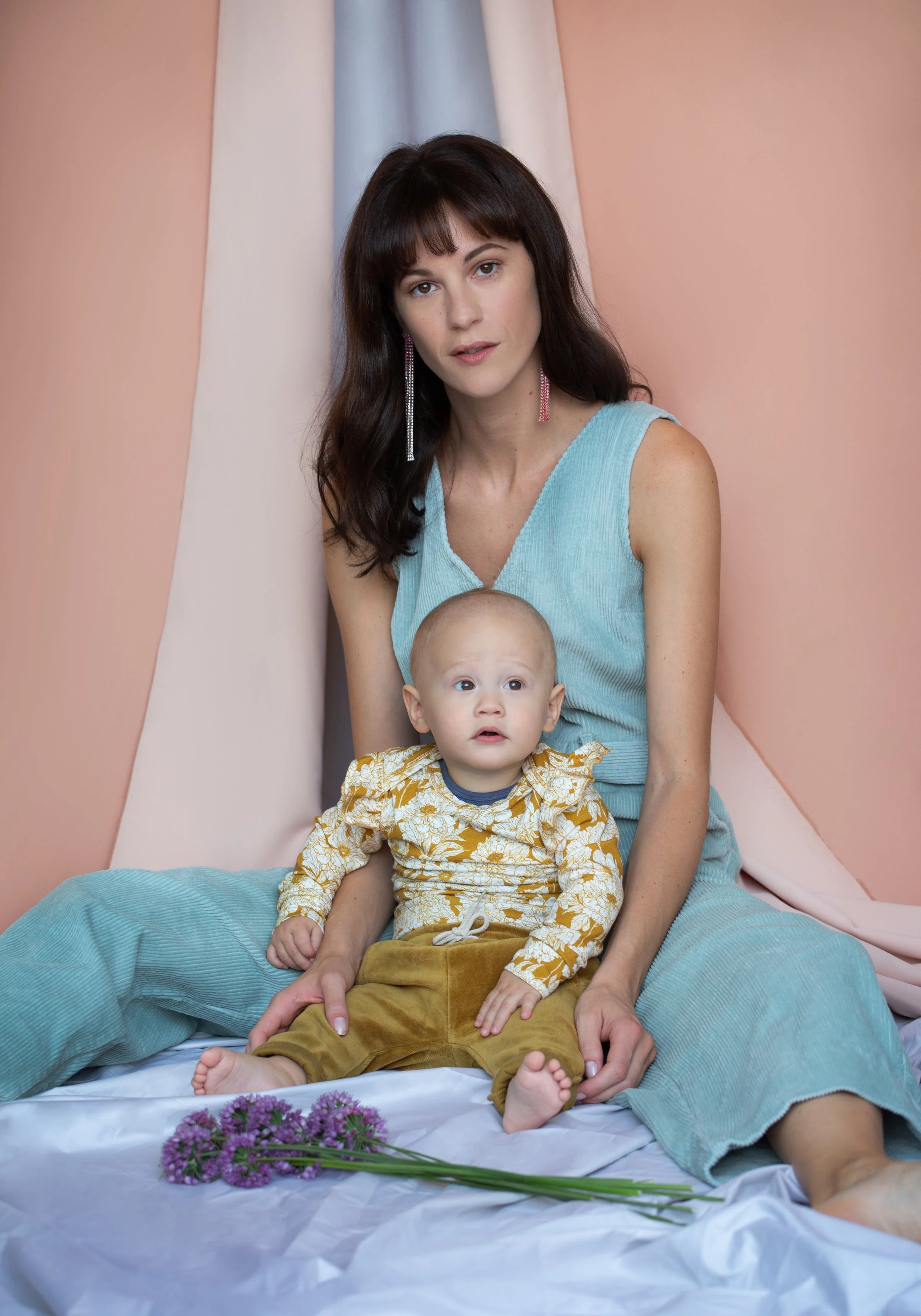 A woman with dark hair and light skin sitting on the floor with a young child on her lap. The woman is wearing a sleeveless blue top and earrings, looking at the camera. The child has light skin, a bald head, and is wearing a yellow floral outfit, al