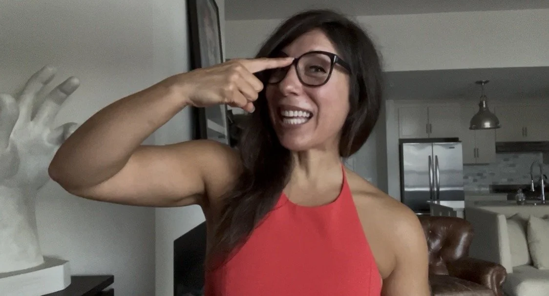 Woman with long hair wearing glasses and a red sleeveless top, smiling and pointing to her glasses inside a home.
