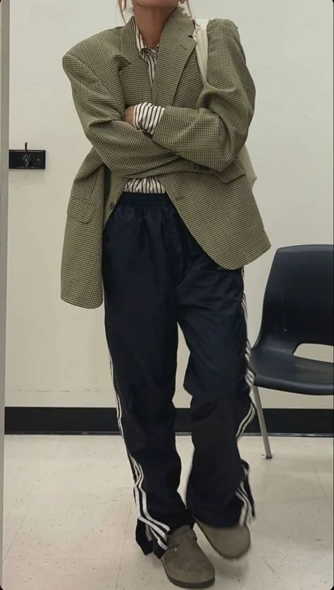 Person wearing oversized blazer, striped shirt, track pants, and loafers; standing indoors next to a chair.