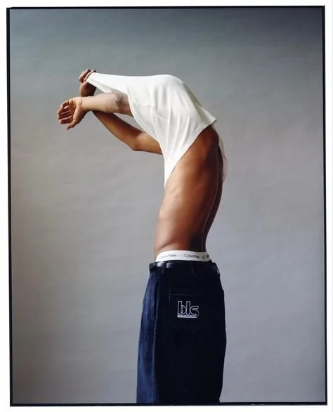 Person removing white shirt, torso visible, wearing Calvin Klein underwear and bls brand pants, against neutral background.