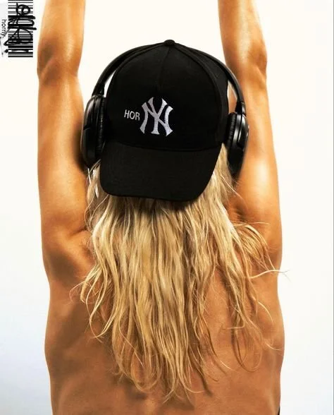 Person with long blonde hair, wearing a black cap and headphones, viewed from the back.