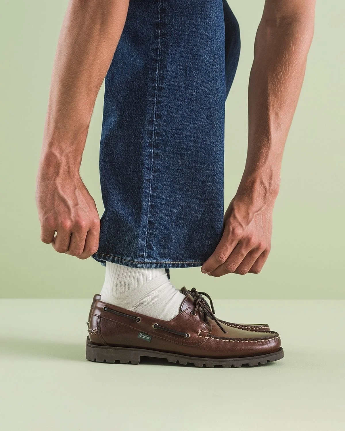 Person in blue jeans and white socks adjusting brown leather shoes against a light green background.