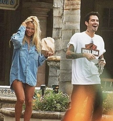 Two people outdoors laughing and holding items; one wearing a blue shirt, the other a white T-shirt.