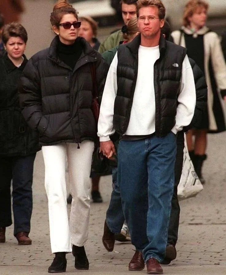 A couple walking outdoors in casual winter clothing, wearing sunglasses and holding hands, surrounded by other people.