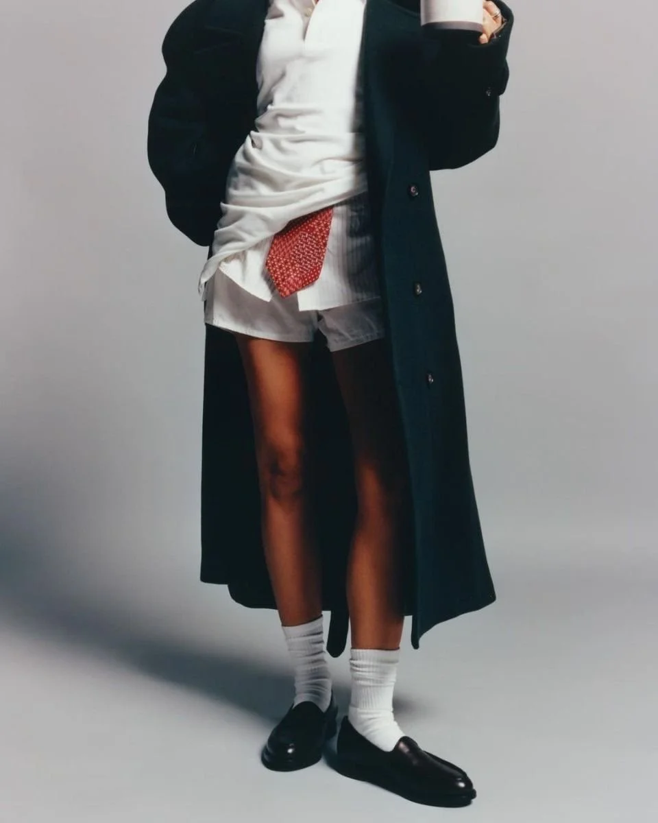 Person dressed in a long dark coat, red patterned tie, white shirt and shorts, wearing white socks and black loafers, holding a mug.