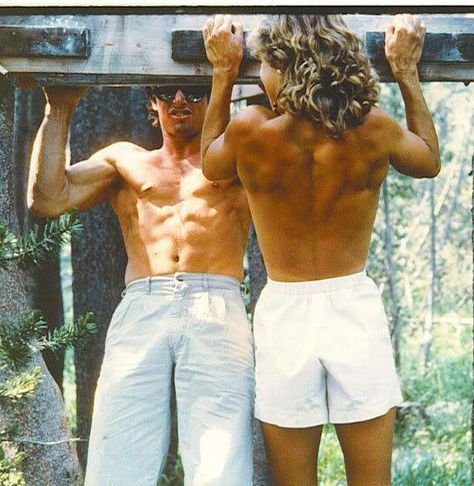 Two shirtless individuals doing pull-ups on an outdoor wooden structure.