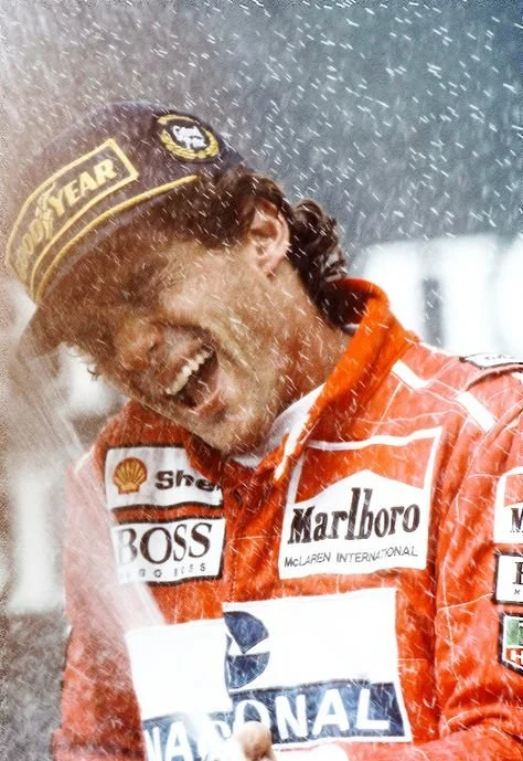 Race car driver celebrating with champagne spray, wearing a red racing suit and cap with sponsor logos.